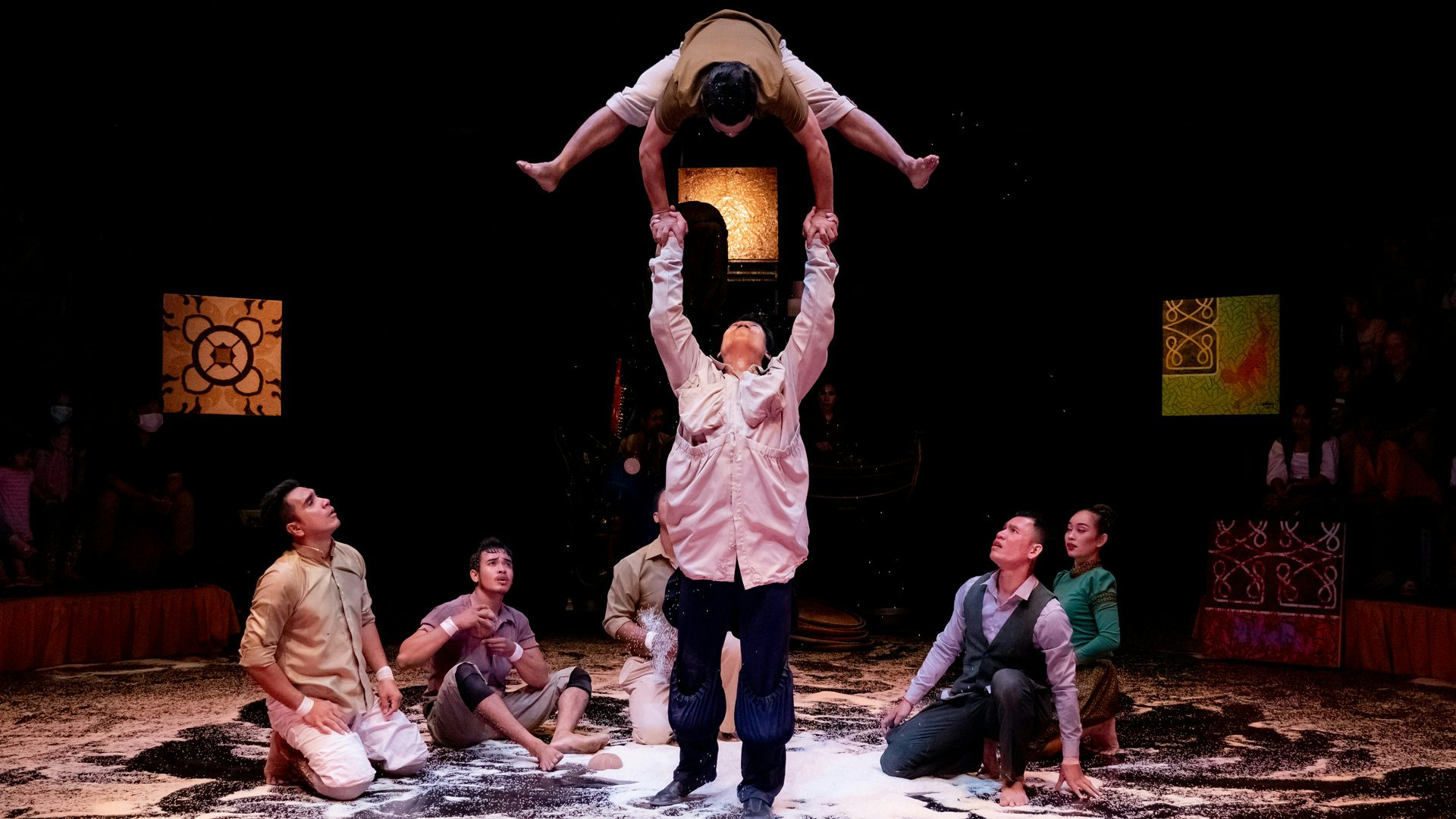 Two people perform acrobatics on a stage covered with white rice as five people look on while kneeling around the acrobats.