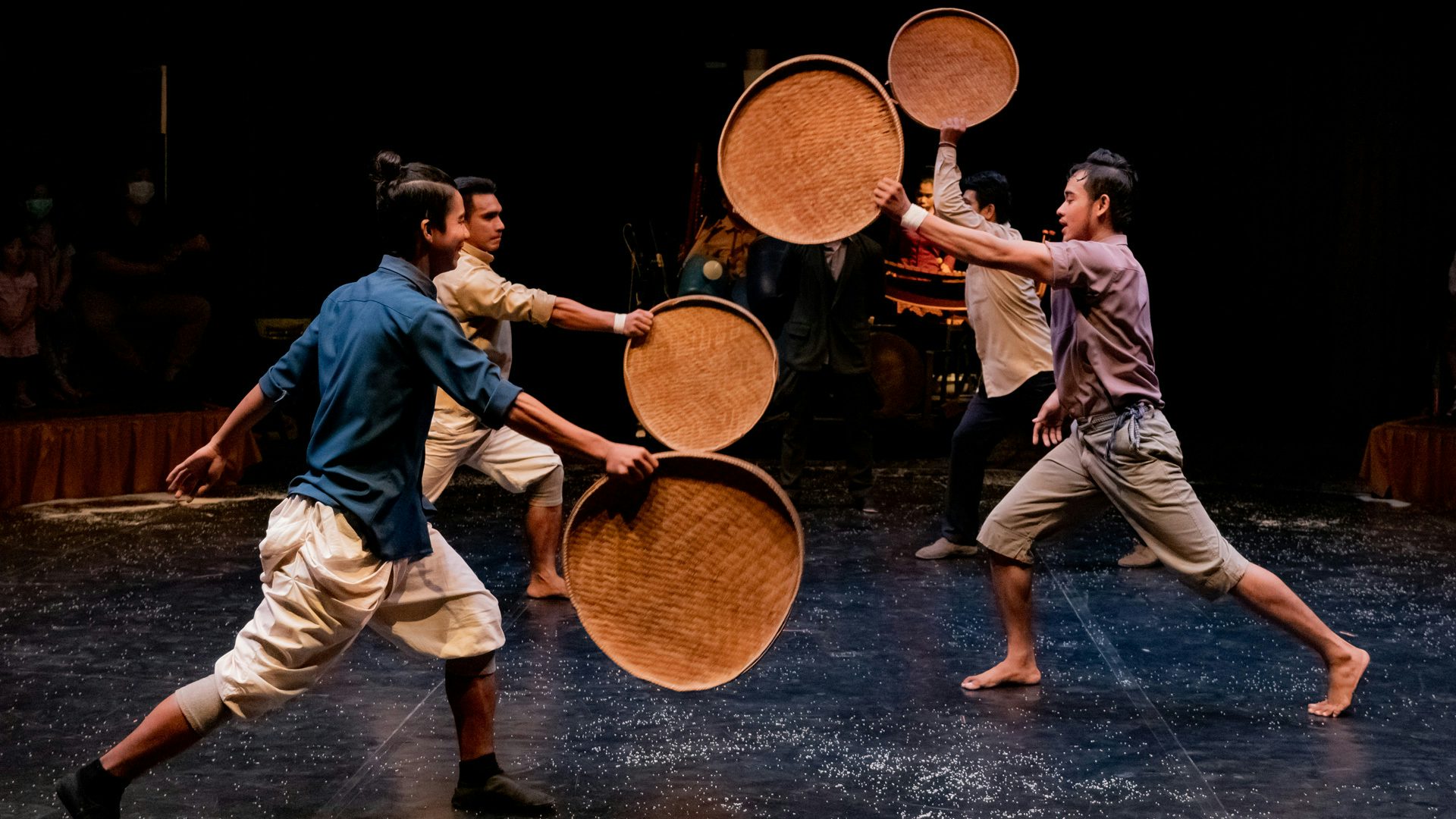 A group of four people dance on stage with woven trays.