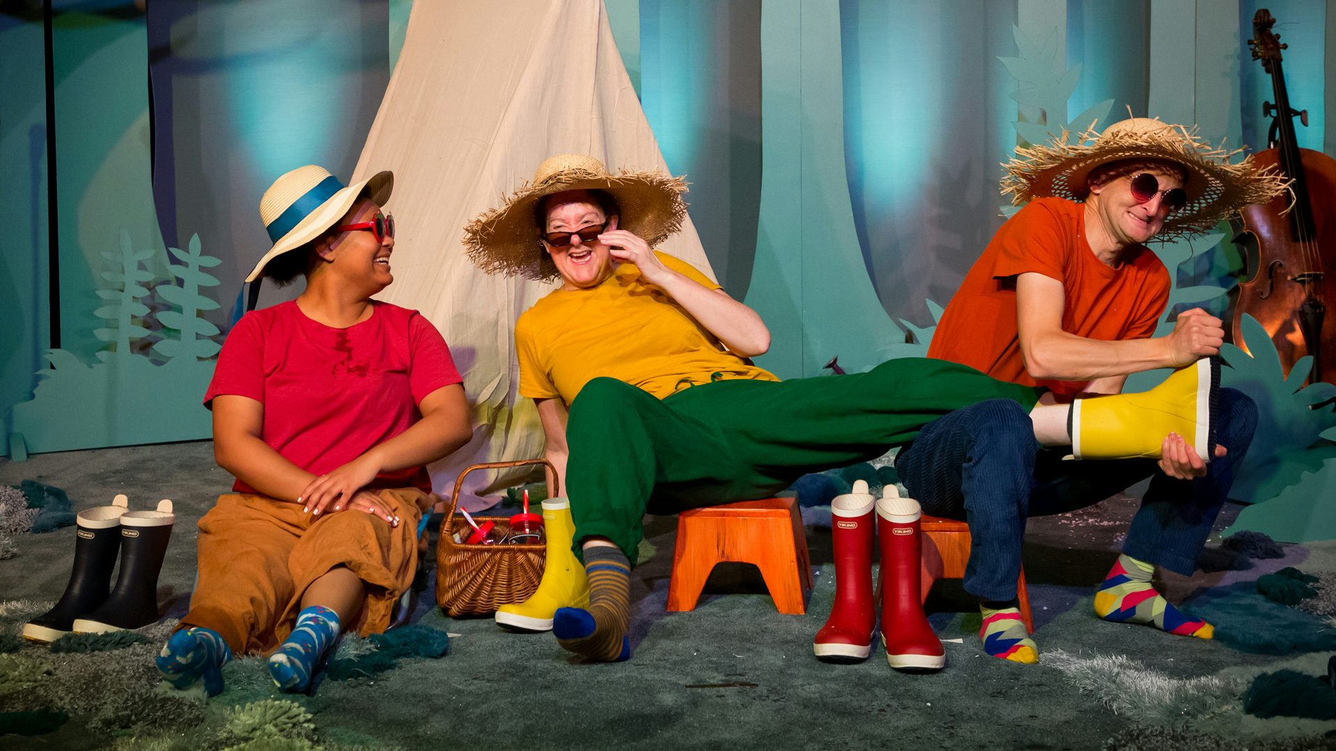 Three people wearing straw hats and sunglasses sit side by side at a campsite. The one in the middle wears a yellow shirt, green pants and a single yellow boot, which the person on the right, wearing a red shirt, is struggling to take off. The person on the left, also wearing a red shirt, looks on and smiles.