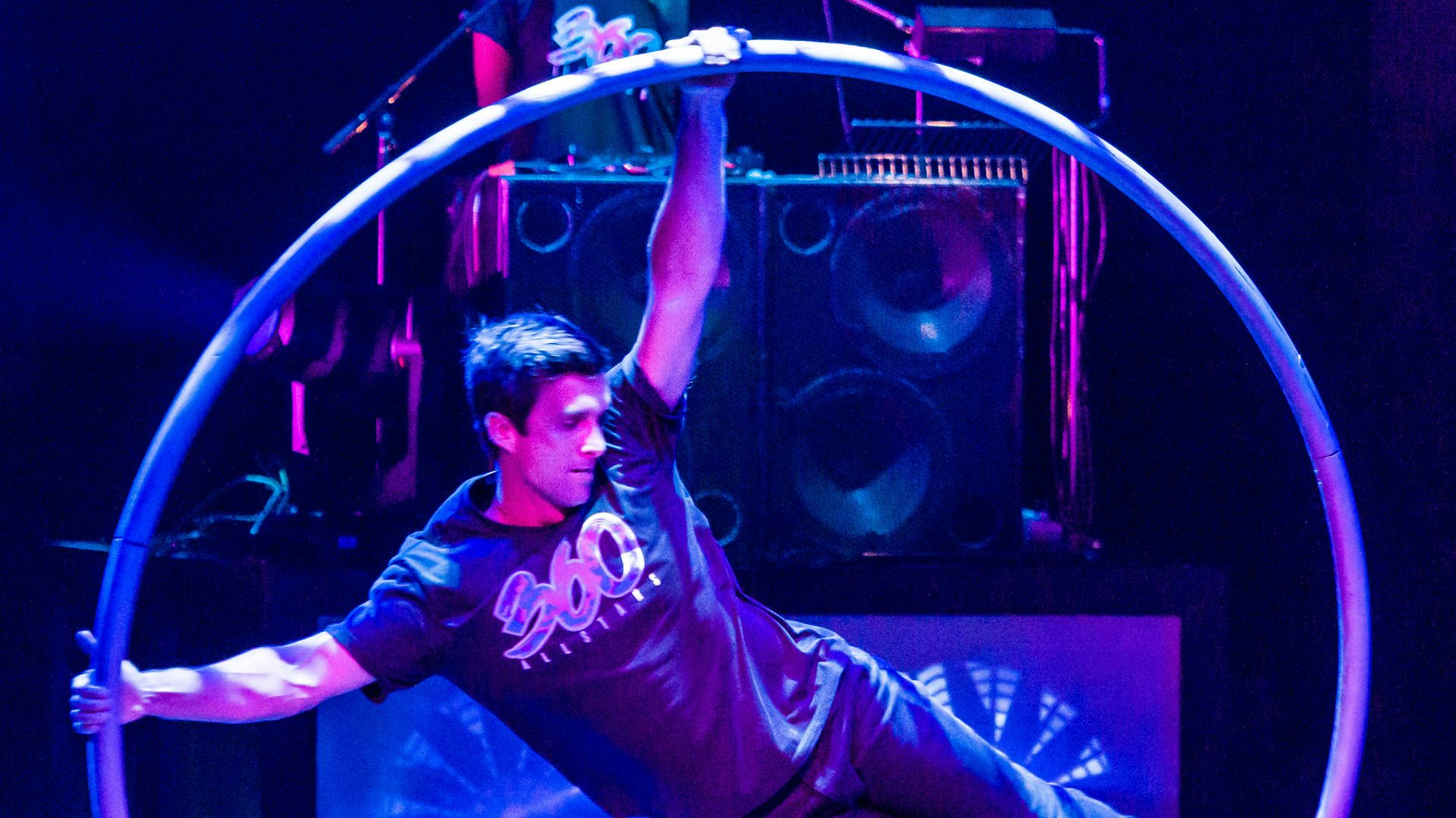A man wearing a black shirt with the show’s name (360 ALLSTARS) does circus tricks in a metal hoop in front of a big amp. Blue and purple lights shine on the stage.
