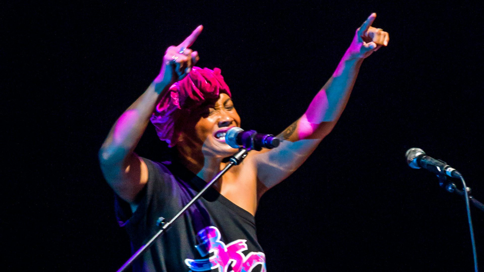 A DJ (disc jockey) talks into a microphone while pointing both hands in the air. The DJ wears a black shirt with the show’s name (360 ALLSTARS) in purple.