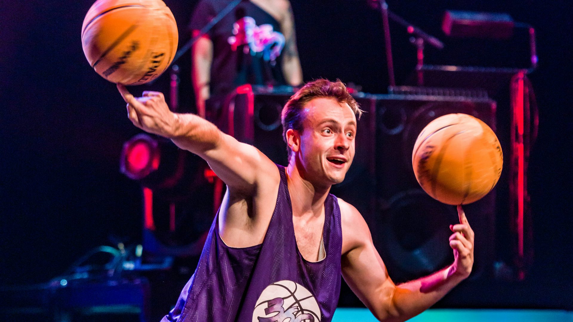 A man spins two basketballs with his fingers on both hands while a DJ stands in the background.