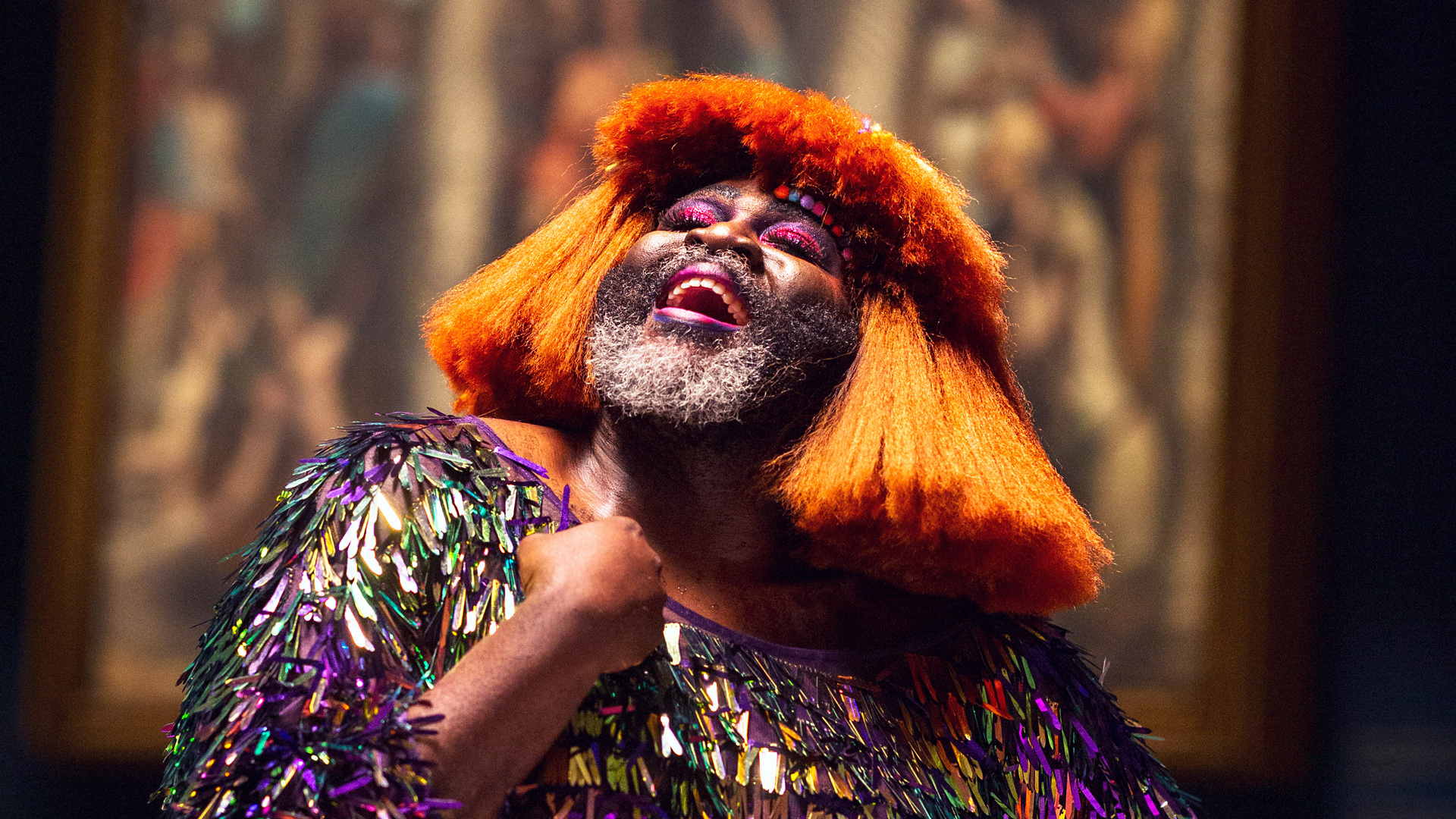 A person with a beard, colorful makeup and short orange hair wearing a colorful, sequined shirt smiles and looks up while holding their hand to their chest
