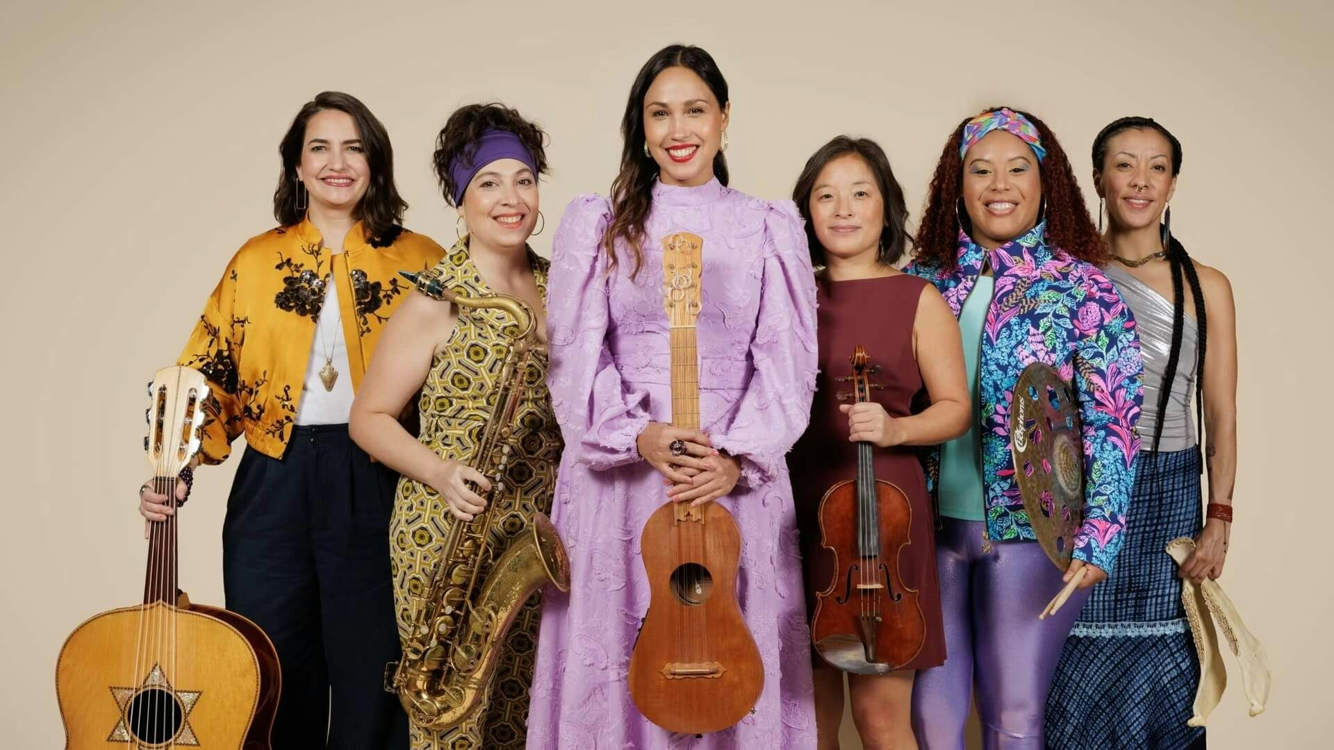 Six women in colorful clothing smile and stand side by side in a line while holding instruments
