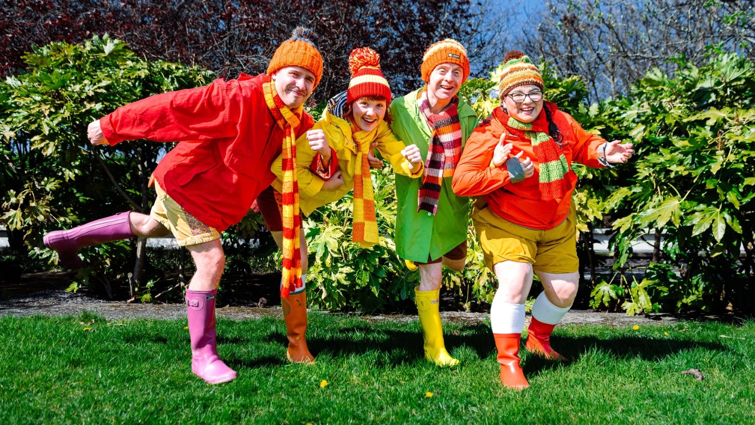 A group of four people wear colorful hats, scarves, jackets, shorts and wellies. They stand arm in arm, with three of them standing on one leg, outside in front of grass and bushes.