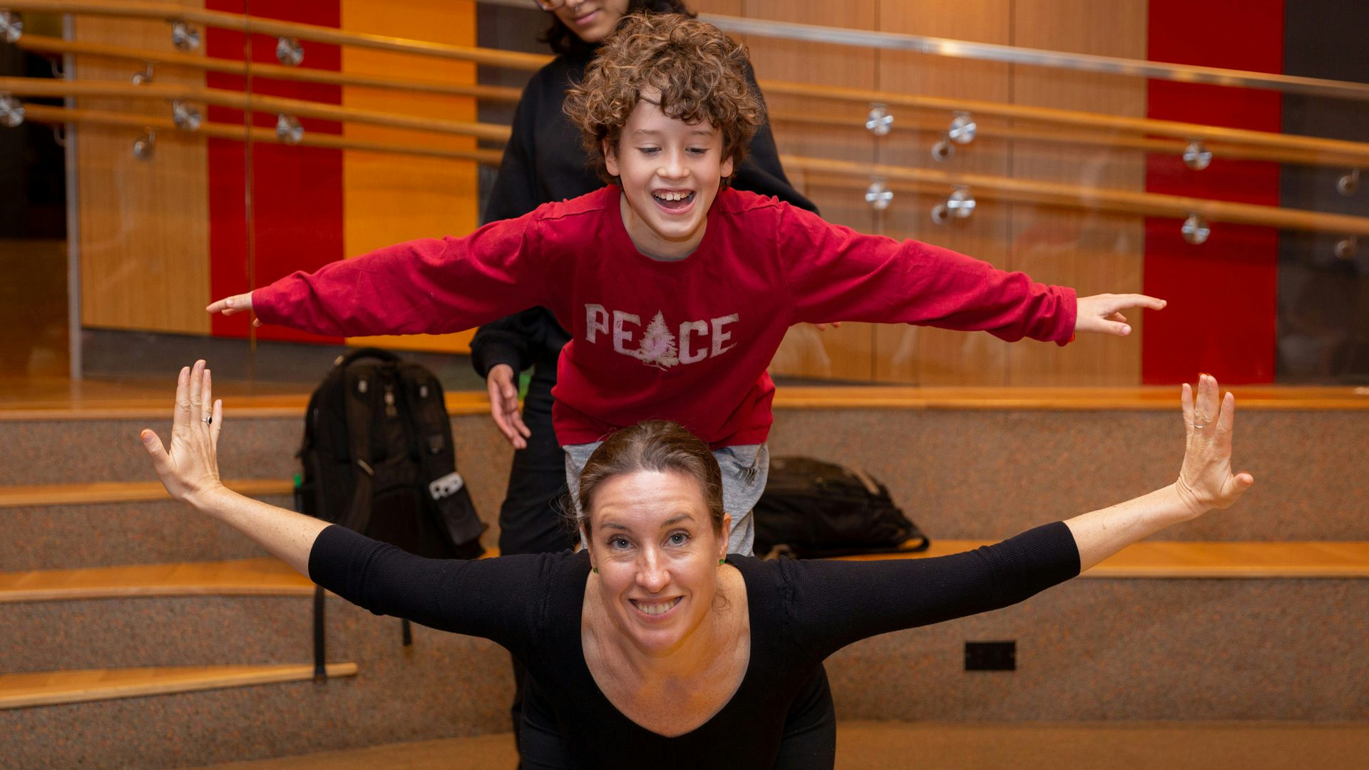 A boy wearing a red shirt balances on his knees while on top of a woman's back. Both have their arms out as if imitating an airplane.