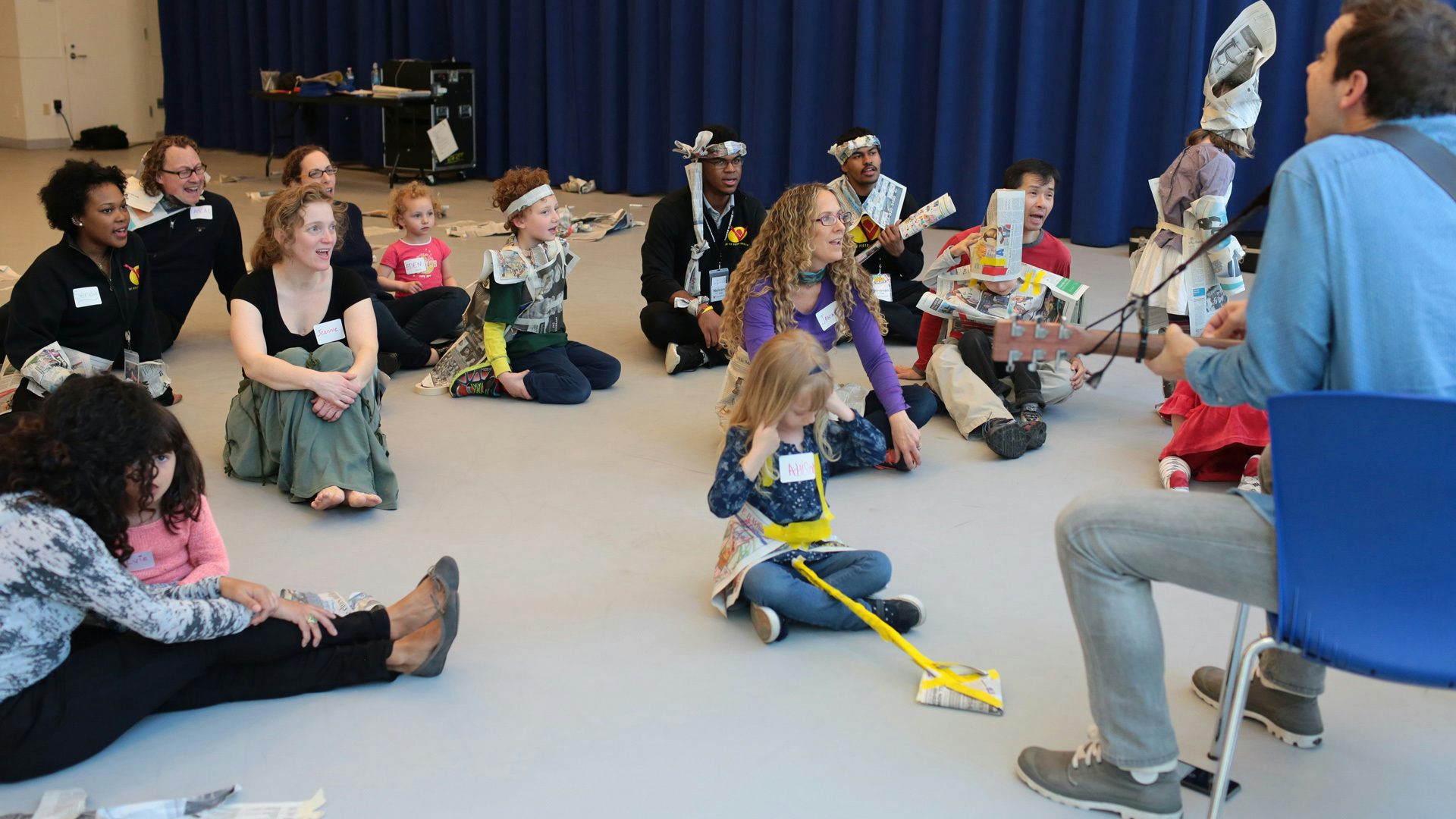 A group of adults and kids, some wearing costumes made out of newspaper, sit around each other as a man sitting in front of the group plays the guitar.