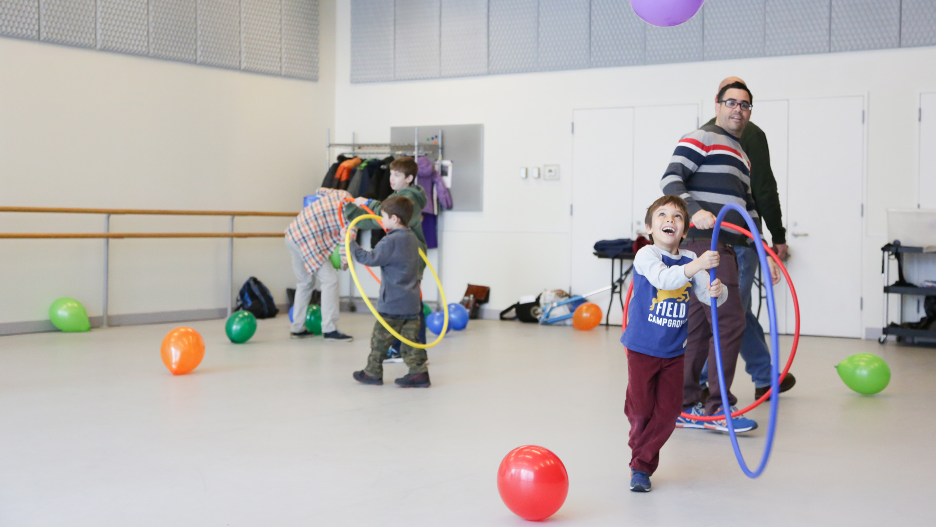 A group of people of varying ages in a room play with multi-colored balloons and hula hoops.