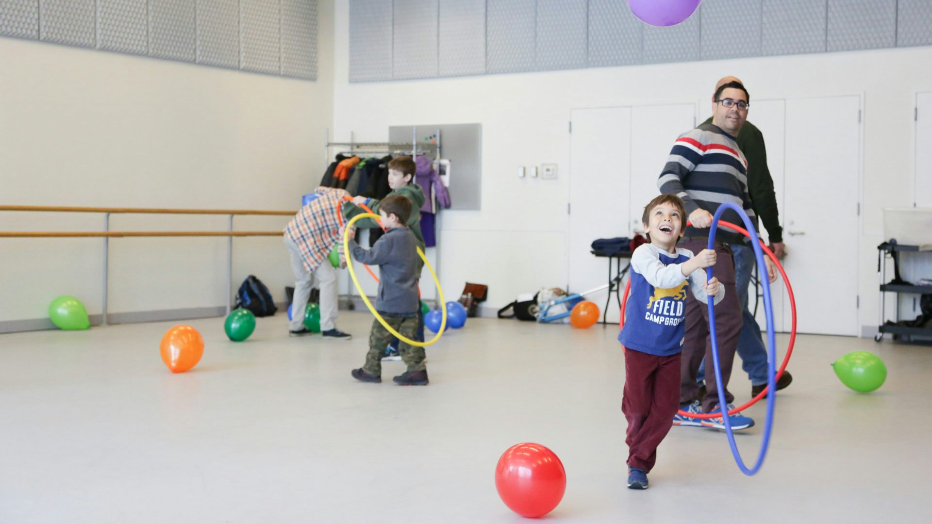 A group of people of varying ages in a room play with multi-colored balloons and hula hoops.