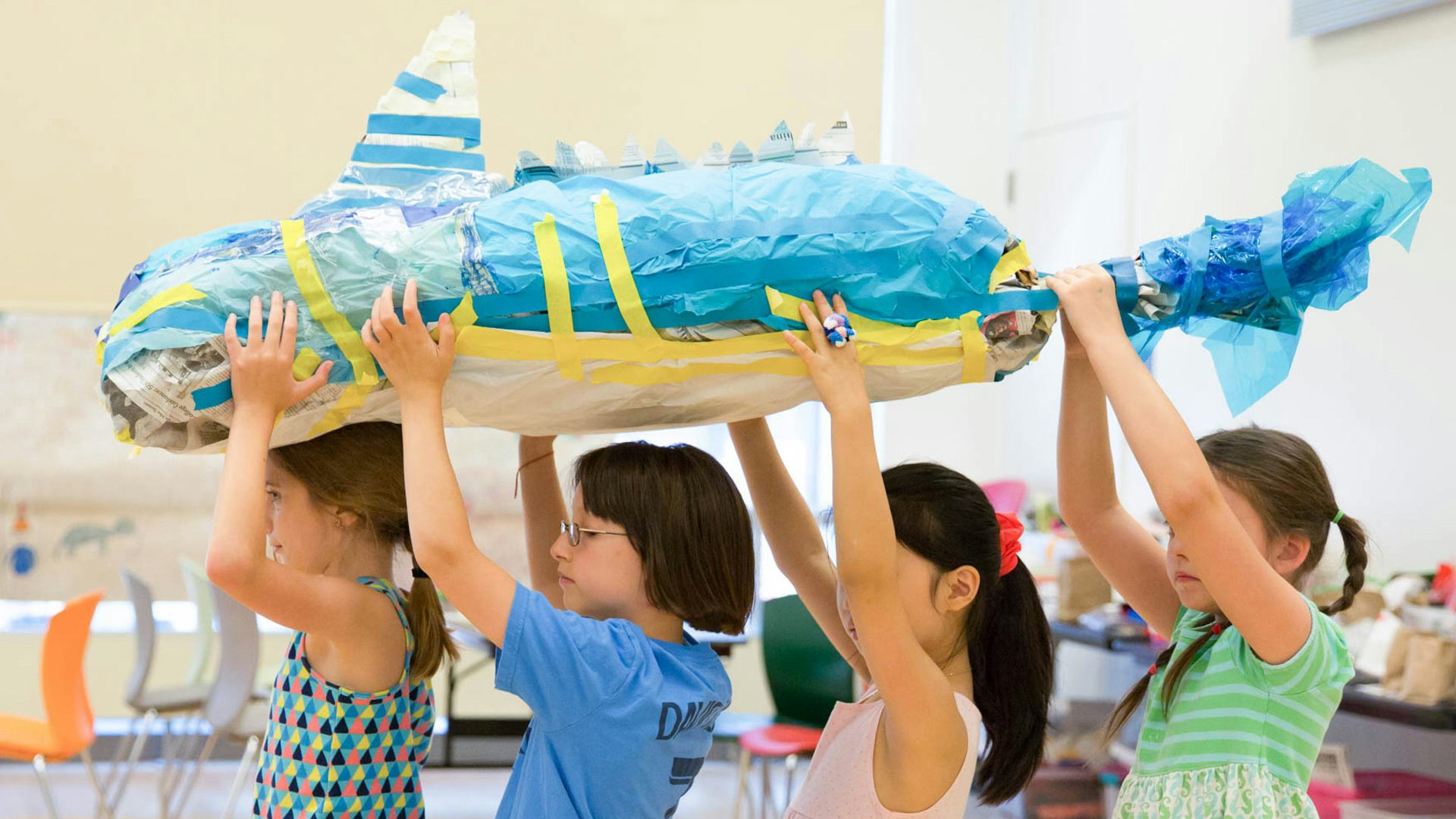 A group of four young children face the left while holding a large, blue, papier-mâché object that has fish/shark-like features above their heads.