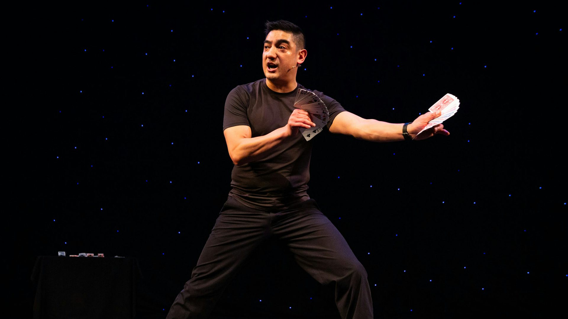 A man in black clothes stands with his legs wide and his arms outstretched as he holds two fanned-out decks of cards in both hands while looking to the left and speaking