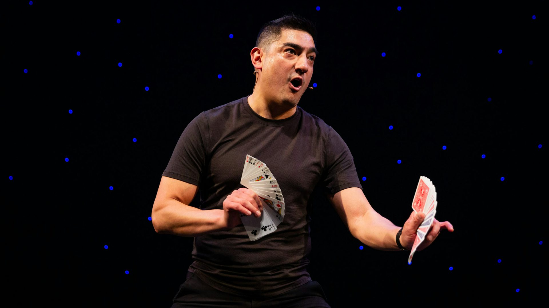 A man in black clothes holds two fanned-out decks of cards in both hands while looking to the right and speaking