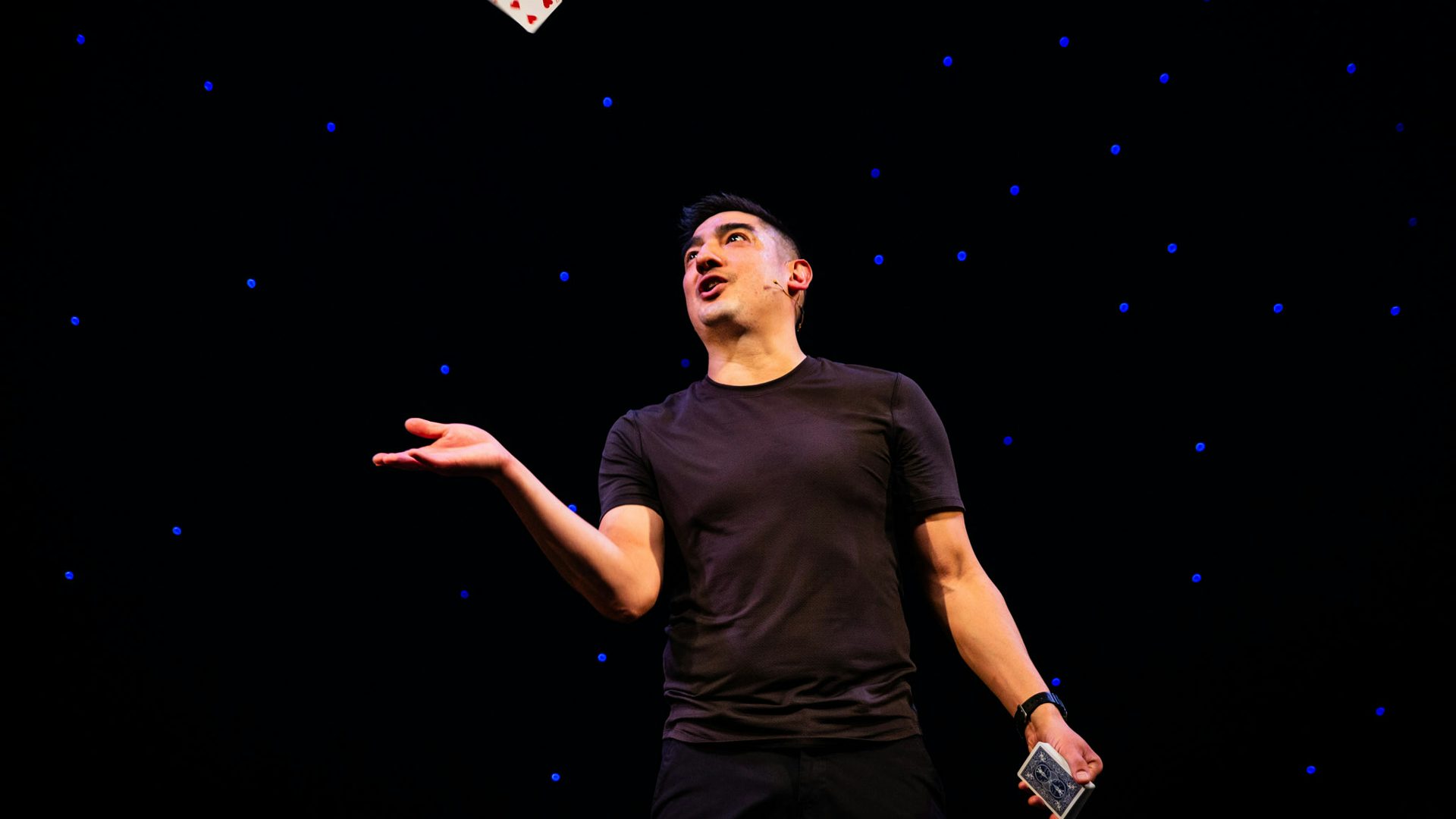 A man in black clothes looks up at a card he threw in the air against a black background with blue dots