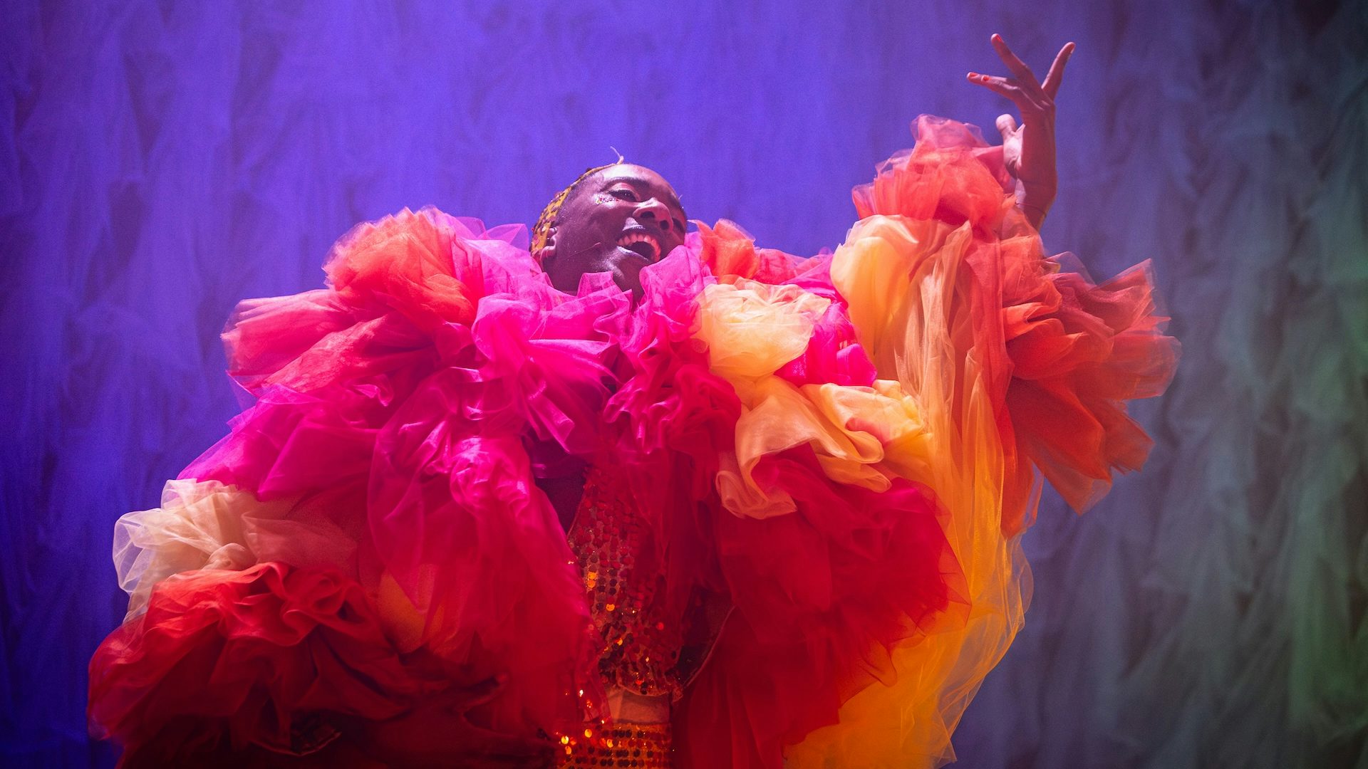 A man wearing large, red and orange ruffles that cover most of his body smiles and holds his left arm up
