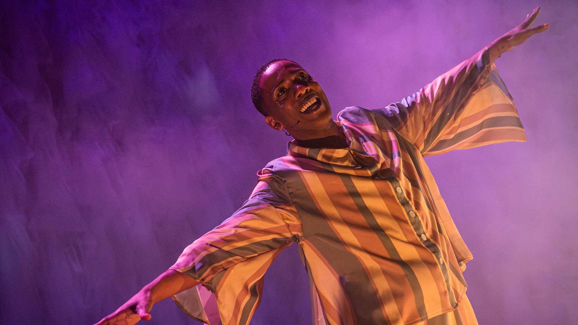 A black man in a tan, brown and white button up shirt smiles and holds his left arm up and his right arm down against a purple background