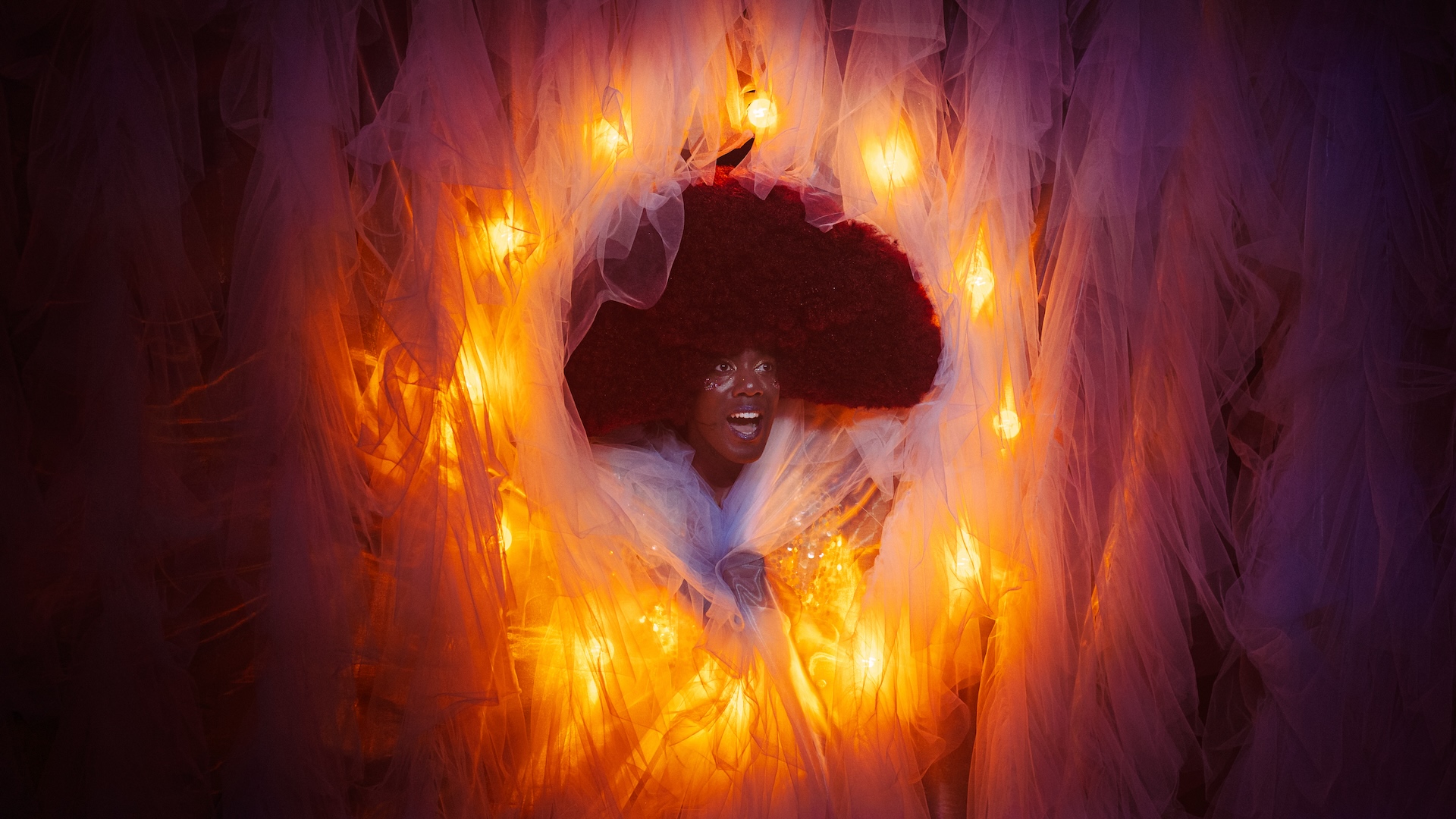 A black man with a large, dark afro and white makeup has a surprised look on his face while peeking out of hole in an orange and purple background