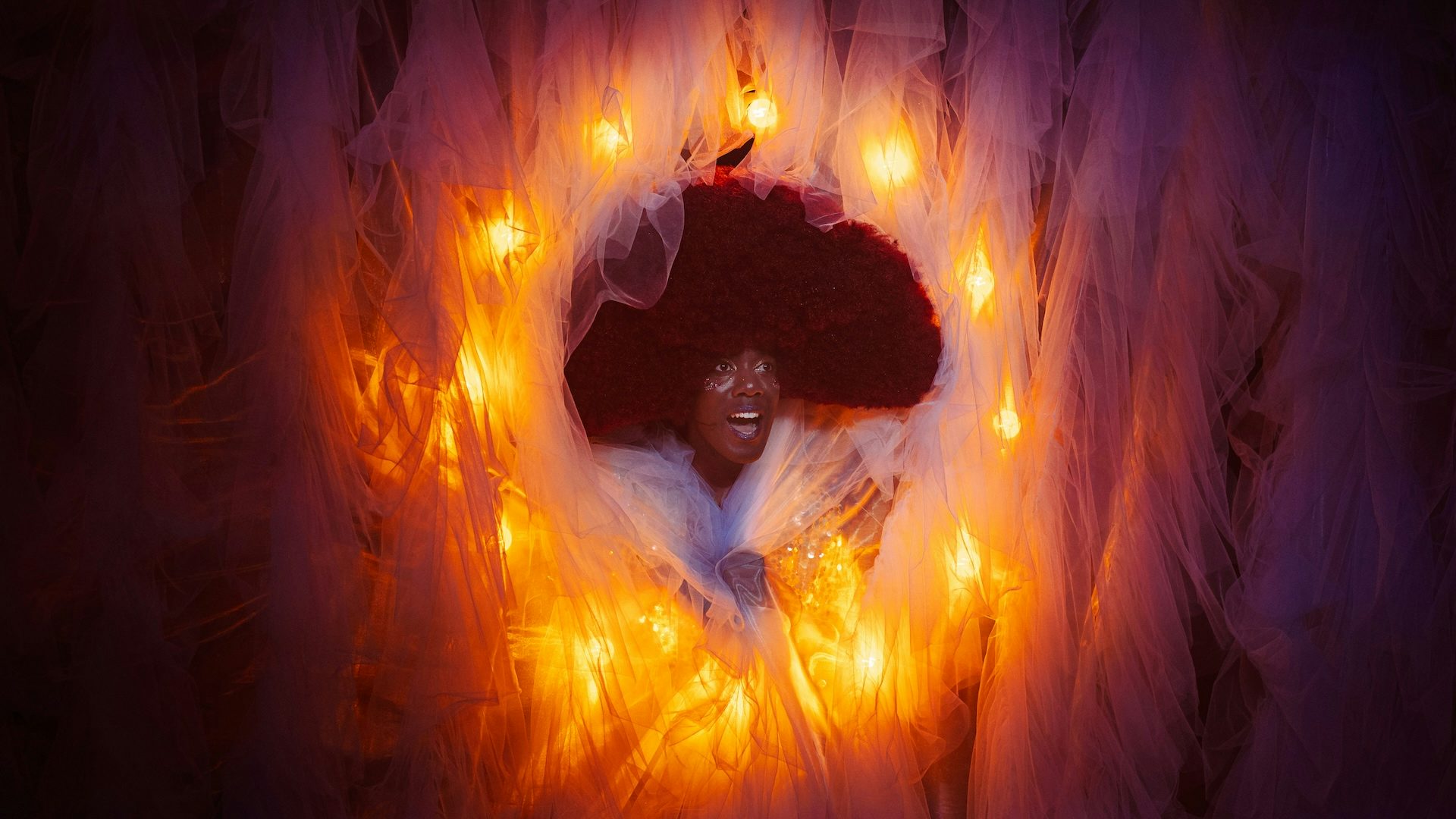 A black man with a large, dark afro and white eye makeup has a surprised look on his face while peeking out of hole in an orange and purple background