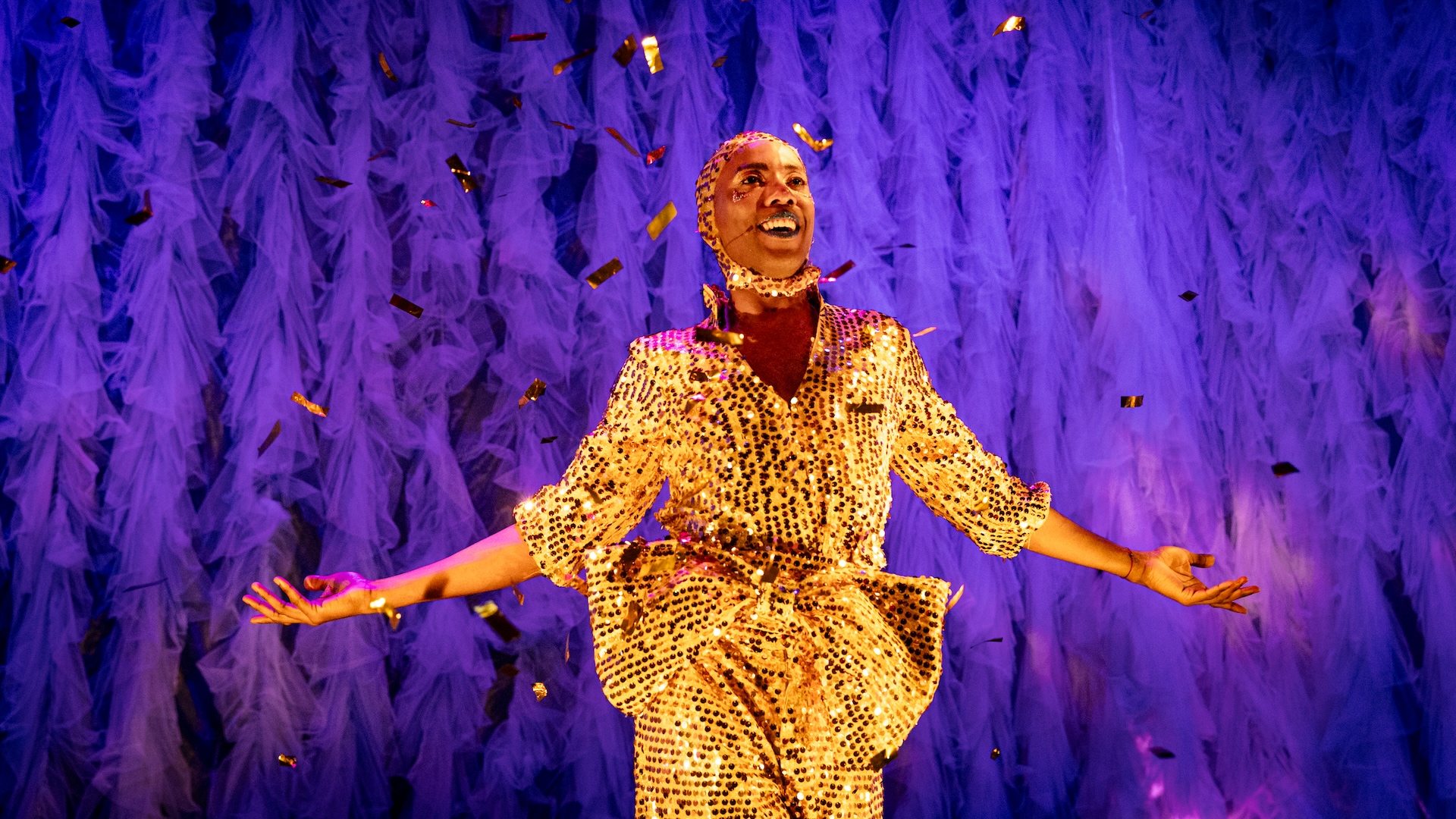 A man in a gold, sequin jumpsuit with a big bow and matching hat stands with his arms out against a purple background