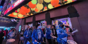 Group of people in a drumline play outside the New Victory Theater beneath the warm, circular lights of the front entrance