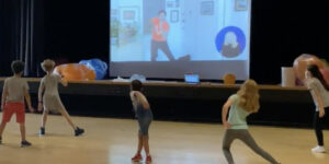 Five kids shown from behind as they follow the movements of a New Victory Teaching Artist wearing an orange shirt on the large screen in front of them