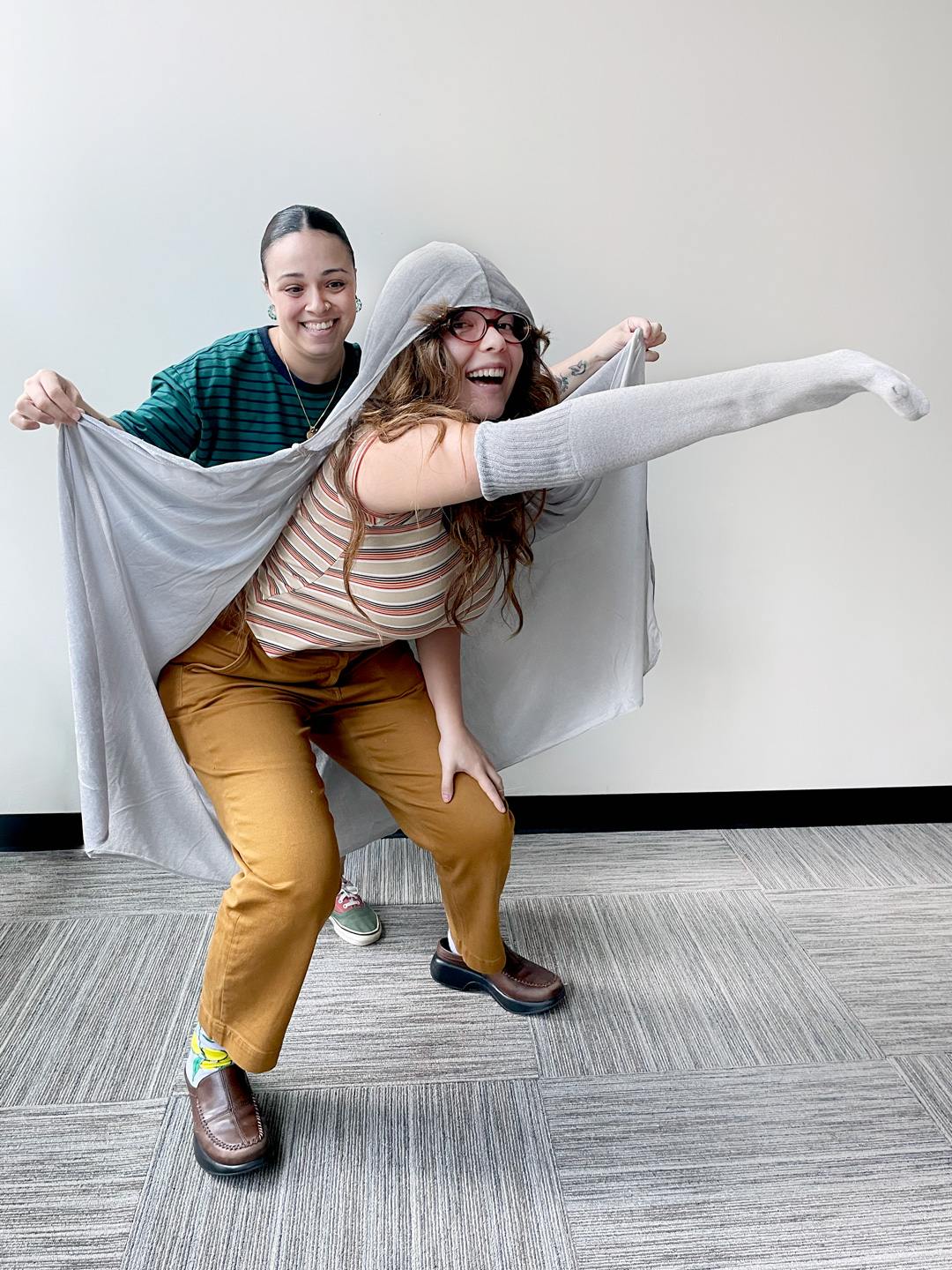 Siobhan pinches the corners of a grey cape that's draped over Kyla's head and shoulders, while Kyla stretches out her sock-wrapped arm like an elephant's trunk