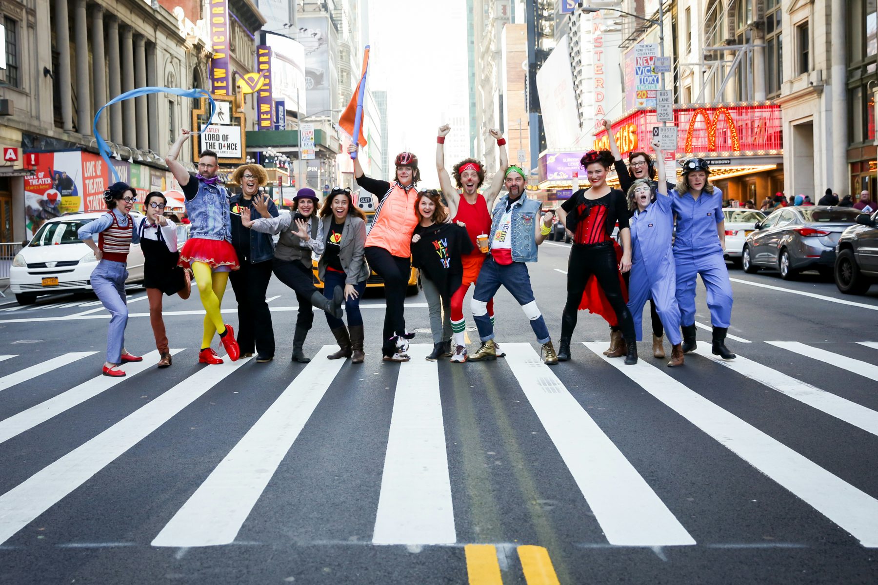 A group of excited people in colorful costumes stand in a line, some with their arms in the air, in the middle of 42nd Street