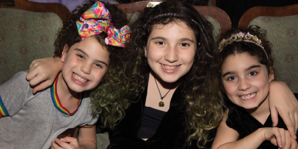 Thee young girls with brown hair and hairbands sitting in theater seats and smiling