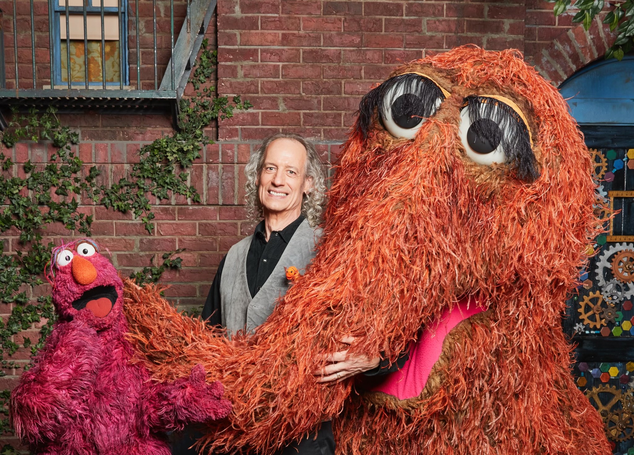 Martin P. Robinson stands between two Muppets: Telly Monster, who is red and furry, and Mr. Snuffleupagus, a woolly mammoth with long eyelashes