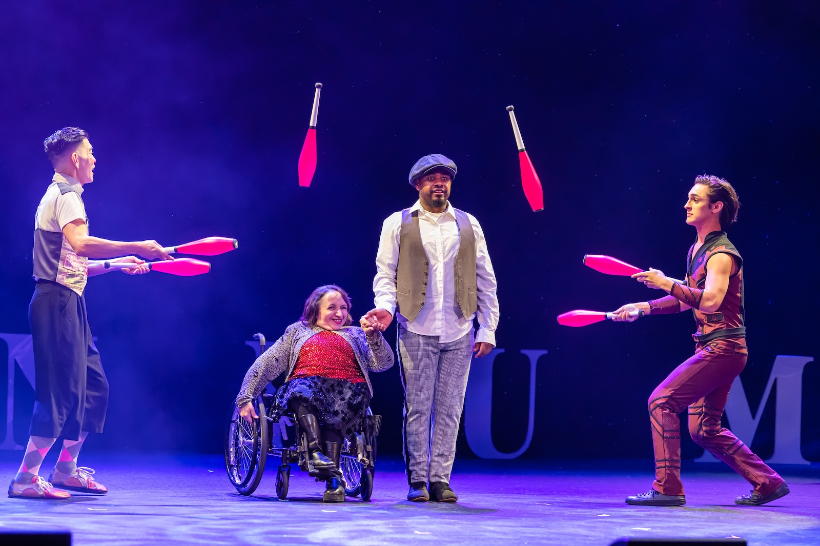 Two jugglers toss juggling pins across the stage, over the heads of a man in a vest and cap and a woman in a wheelchair.