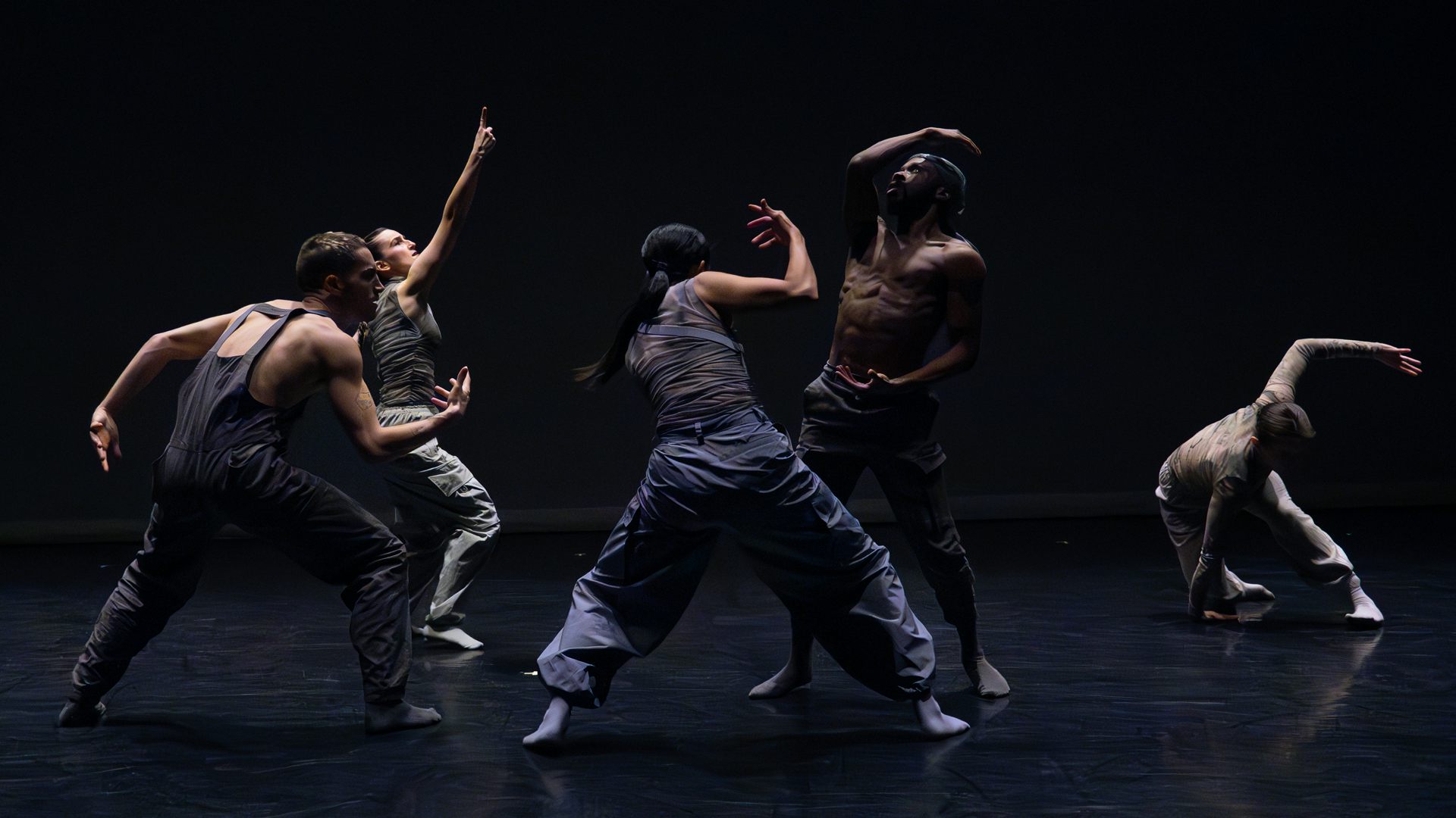 Five dancers in dark clothes stretch their arms out while facing each other against a black background