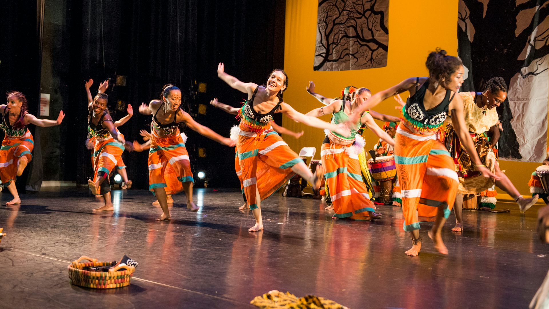 A large group of dancers wearing long, orange skirts with white and green stripes on them lift their left legs and hold their arms out
