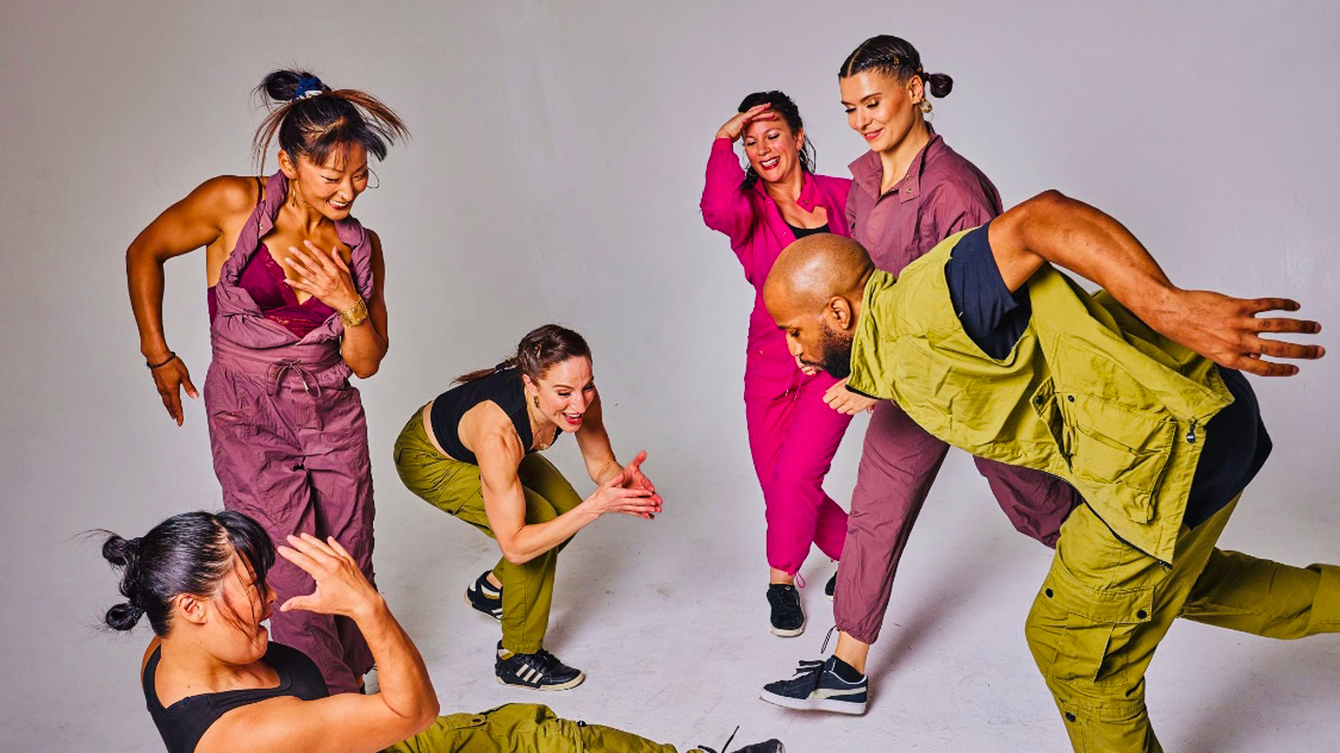 Six dancers in purple, pink and green jumpsuits circle around each other against a gray background
