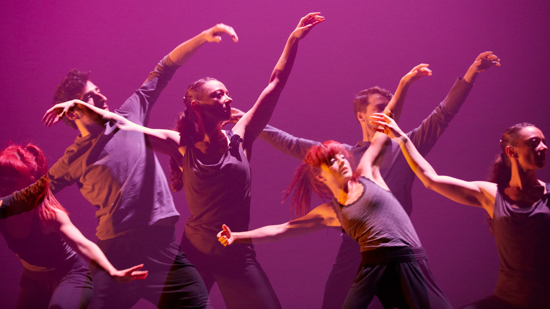 Six dancers in gray shirts and black pants hold their arms up in an "L" shape against a purple background
