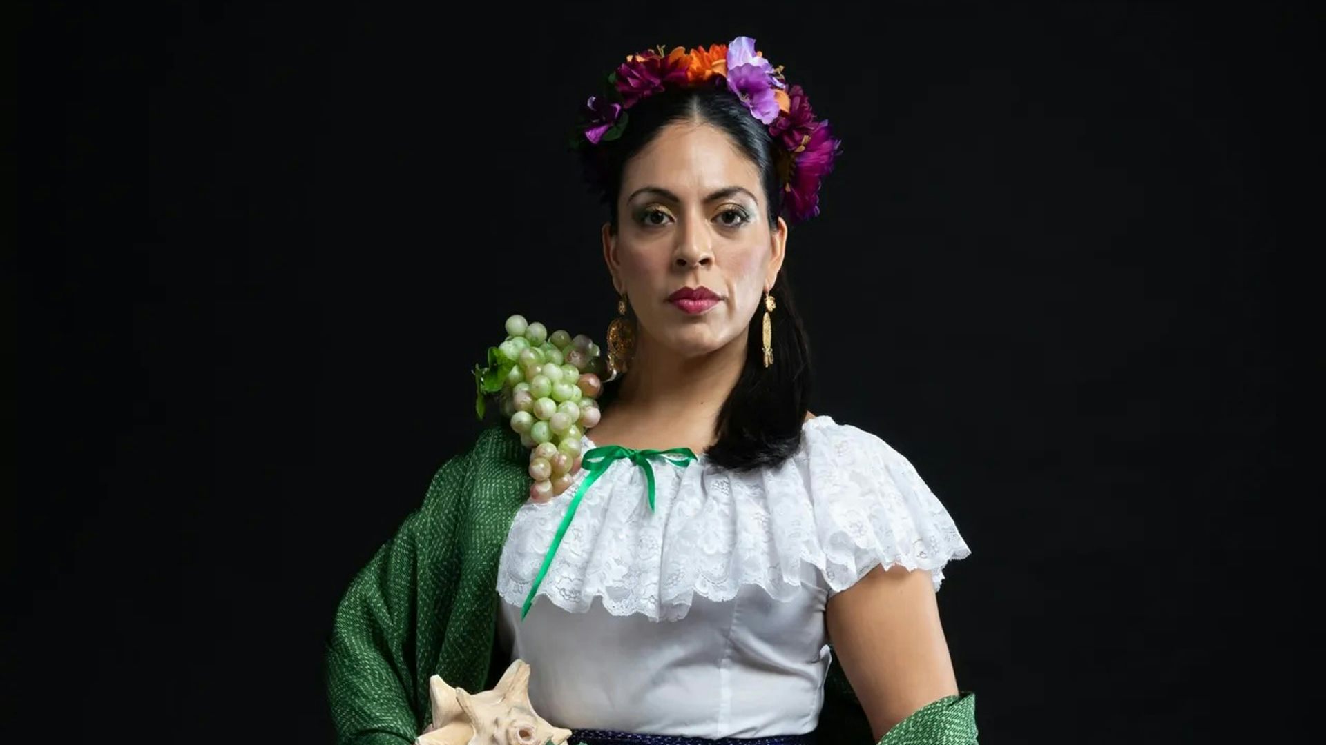 A person with dark hair wears a colorful flower crown, gold earrings, green grapes on their right shoulder and a white blouse while looking straight ahead