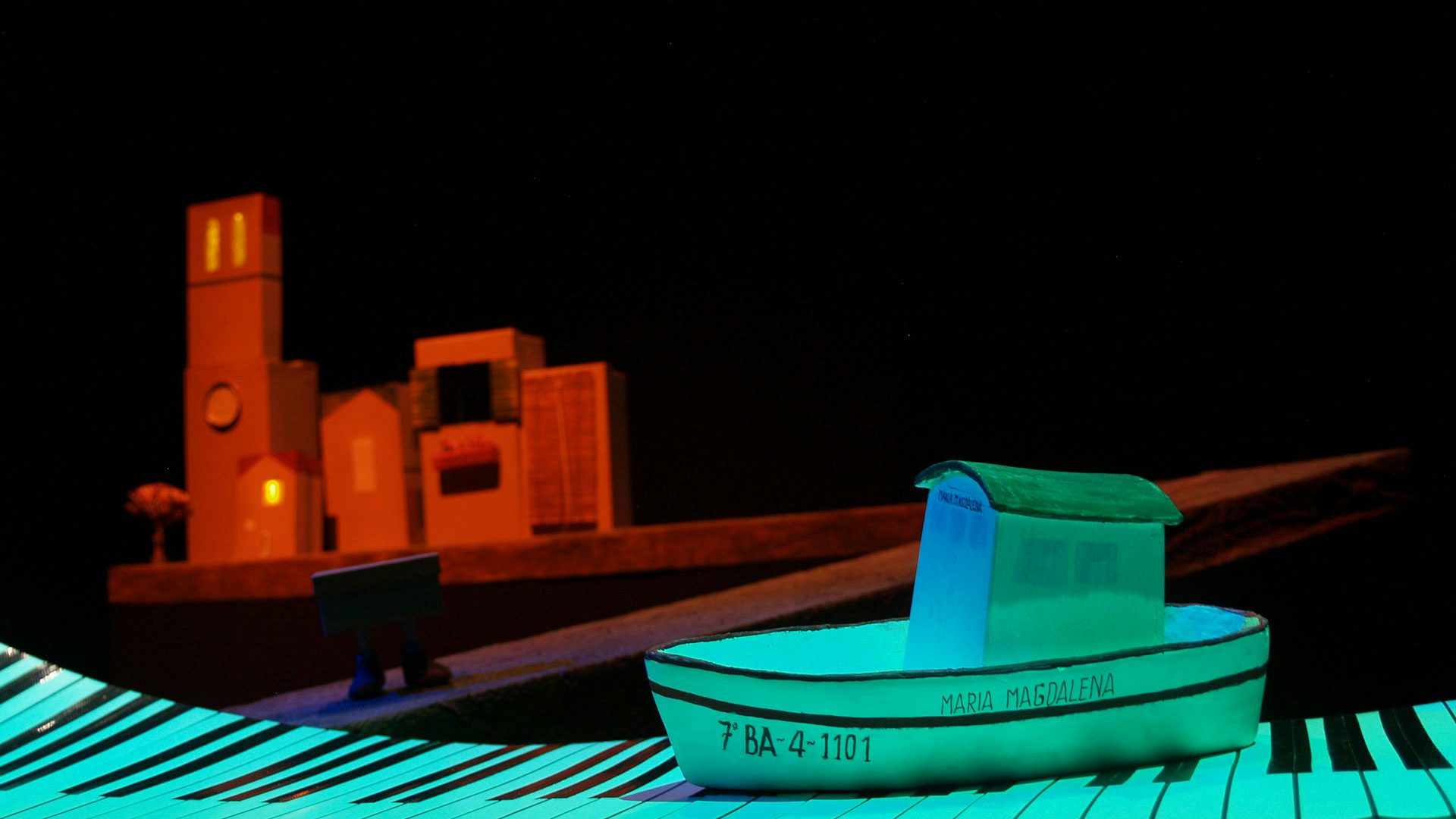 A small prop boat called the Maria Magdalena sits on a piece of paper made to look like piano keys in blue light while a street made out of cardboard boxes is lit in red behind.