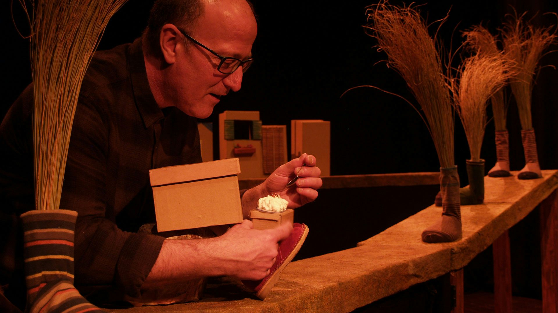 A man with glasses feeds a small prop piece of cake to a puppet of a shoebox that sits on a path filled with various socks that have grain coming out of the top of them.
