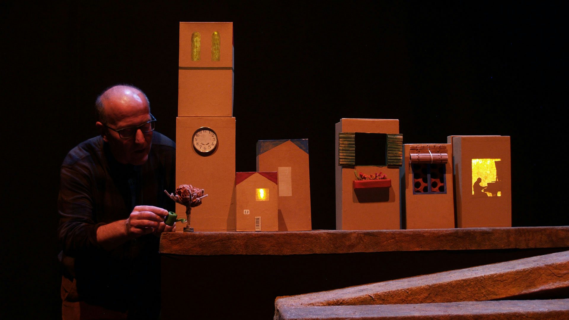 A man with glasses holds a tiny watering can to water a tiny tree on a street built out of cardboard boxes.