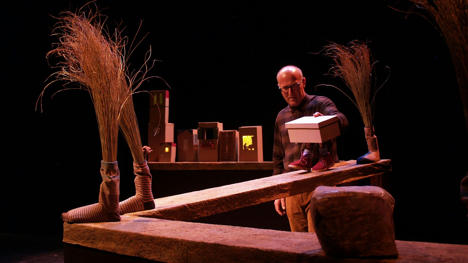A man with glasses puppeteers a shoebox with ankles and red shoes down a zig zag ramp. The ramp also has large socks on it with grain coming out of the top of them.