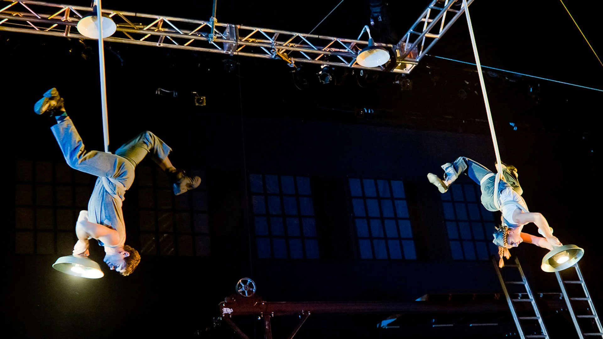 Two performers have their legs outstretched in the air and a hanging light wrapped around their waists.