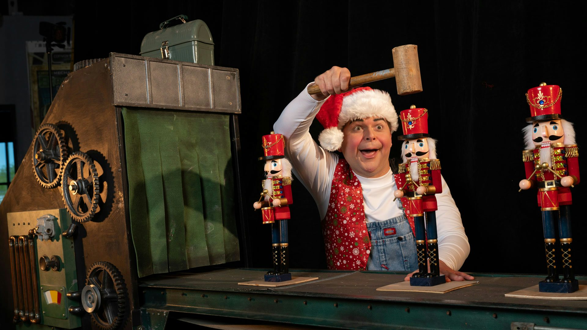 A performer in a Santa hat cheerfully holds a mallet over the middle of three nutcrackers on a mechanical conveyor belt.