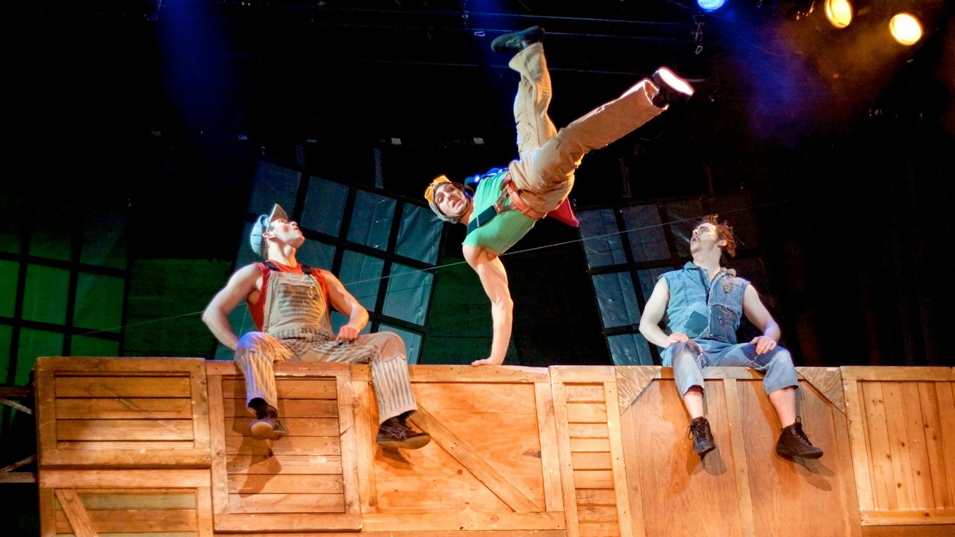 Two performers look in awe toward a third performer doing a funky twisted jump in between them atop a set of boxes.