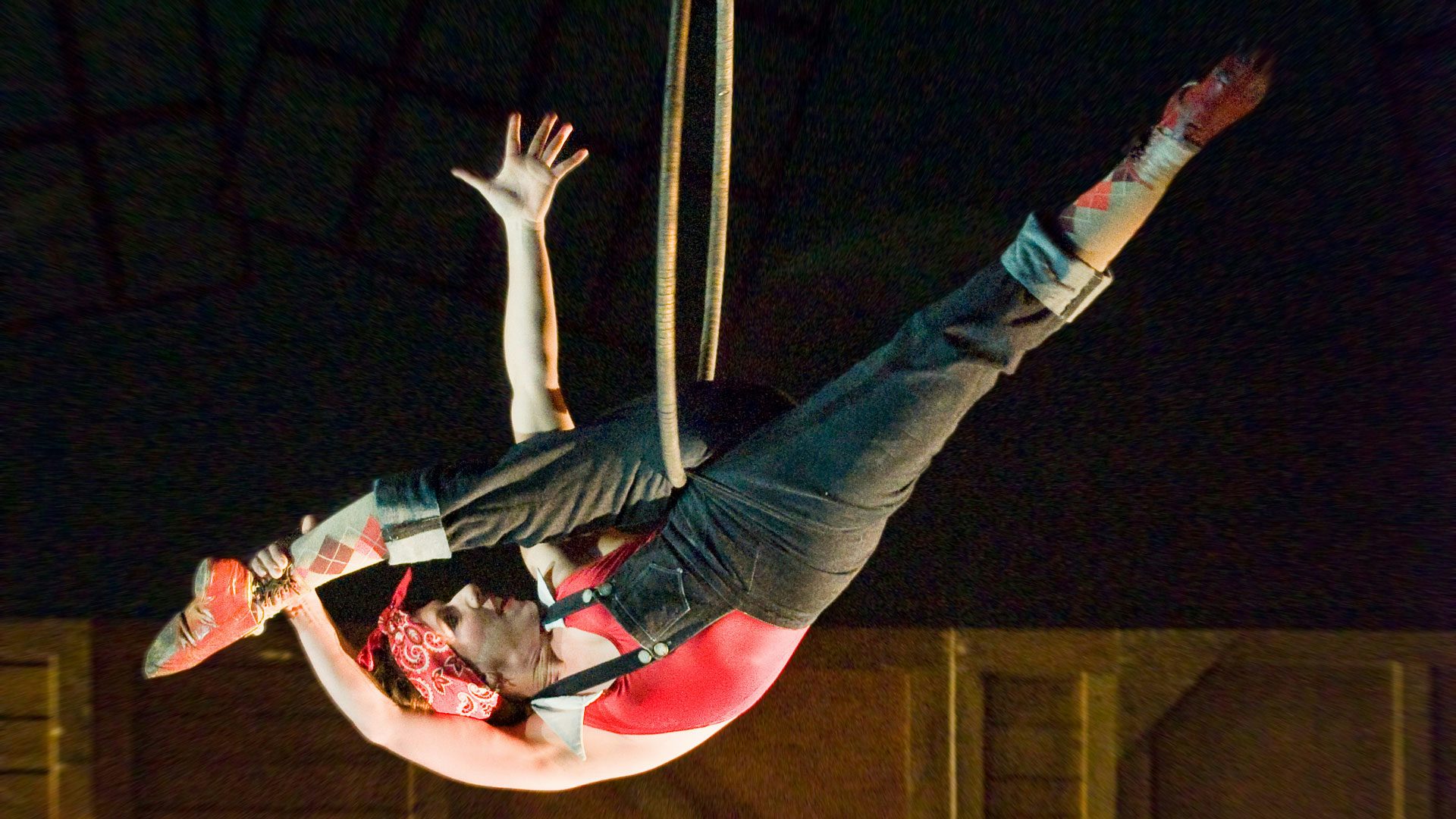 A performer in black overalls, a red bandana and argyle socks stretches their right leg through a rope in a hanging, upside down split.