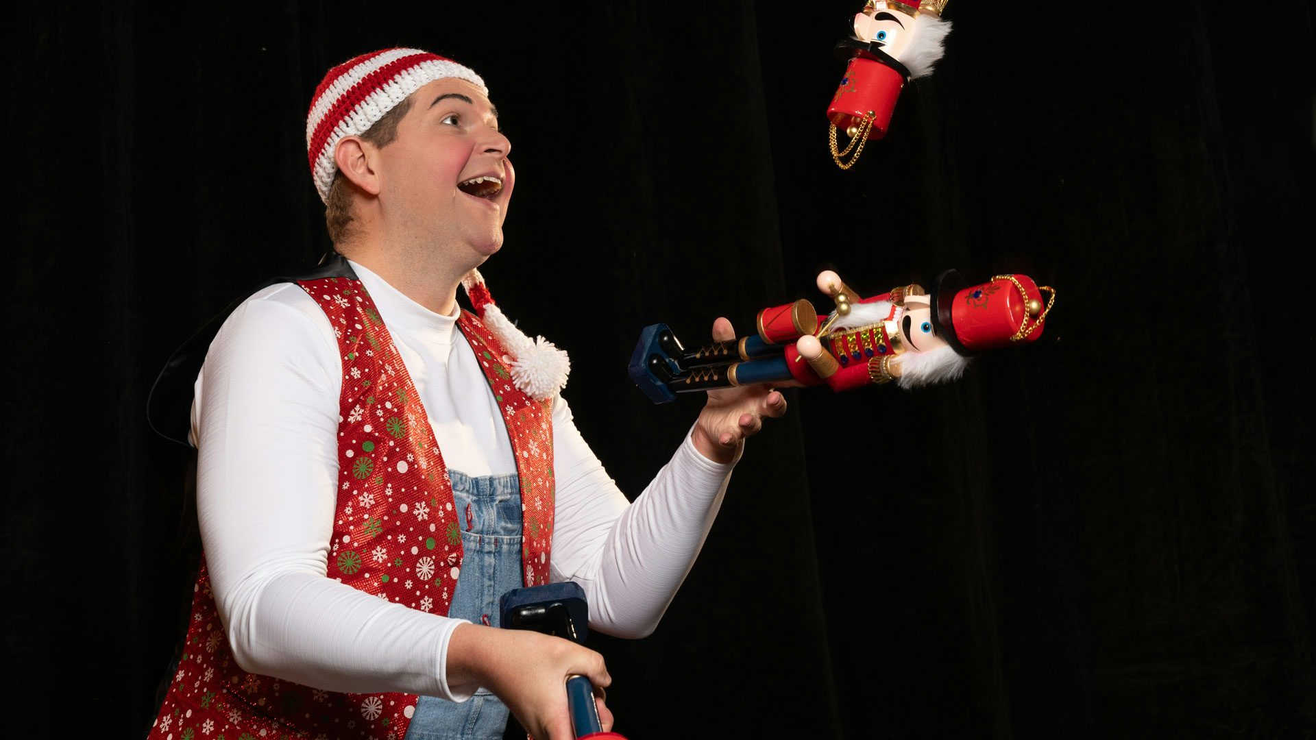 A performer in overalls, a long sleeve white shirt, and a red, white and green vest with a red and white striped hat on juggles three nutcrackers.