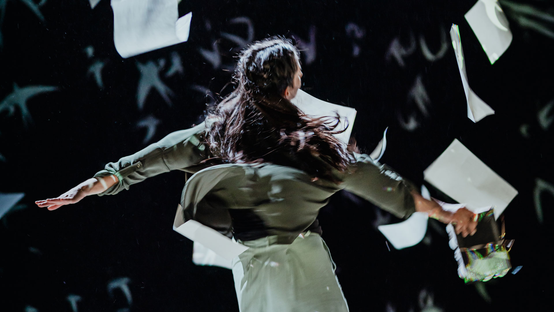 A performer with long hair wearing a long sleeve olive green top and beige pants faces away from the camera, holds a piece of paper and twirls as papers fall around them.
