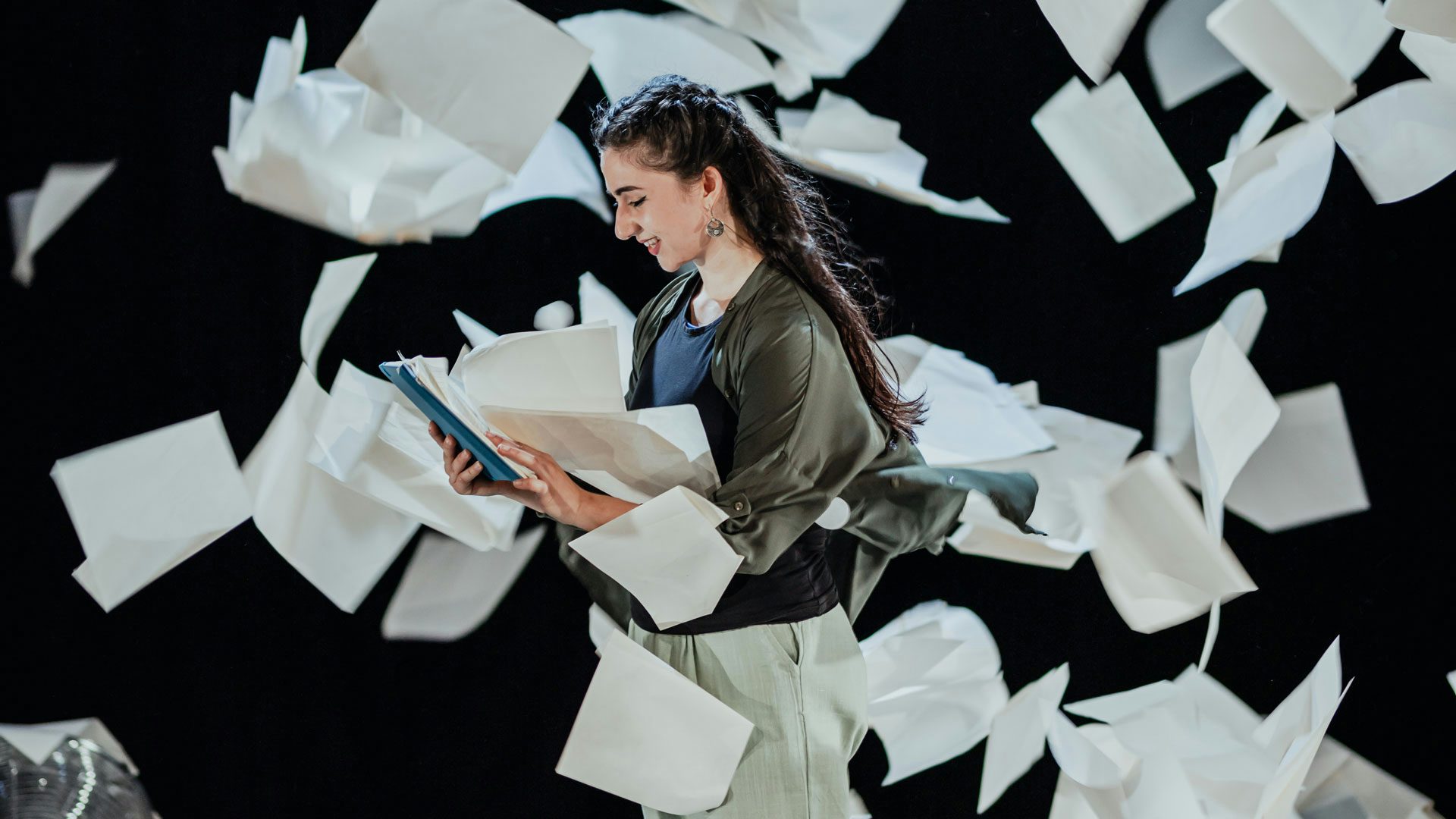A performer with long hair wearing an olive green jacket, navy blue shirt and beige pants smiles at a blue notebook in their hands while papers fly around them.