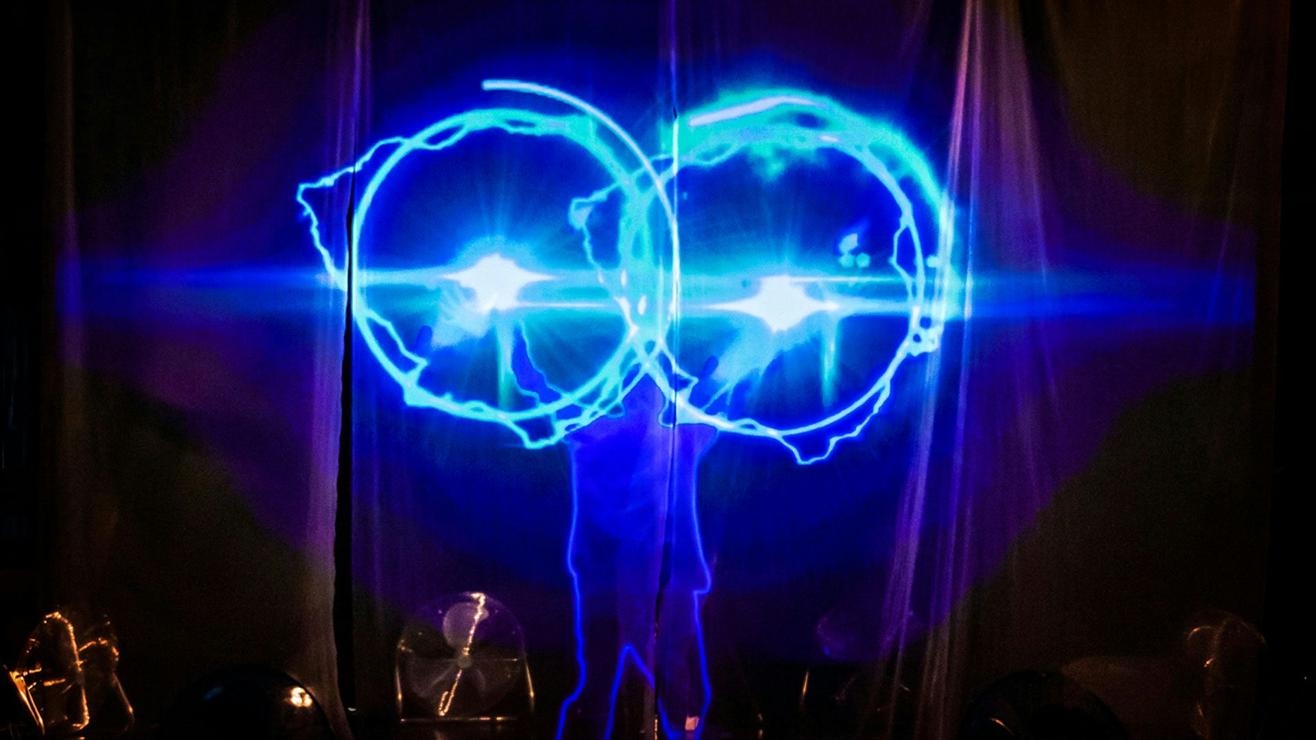 A projection of a performer outlined in blue is against a curtain. The performer holds up their hands and two bright lights appear with two large rings of blue projection surrounding them.