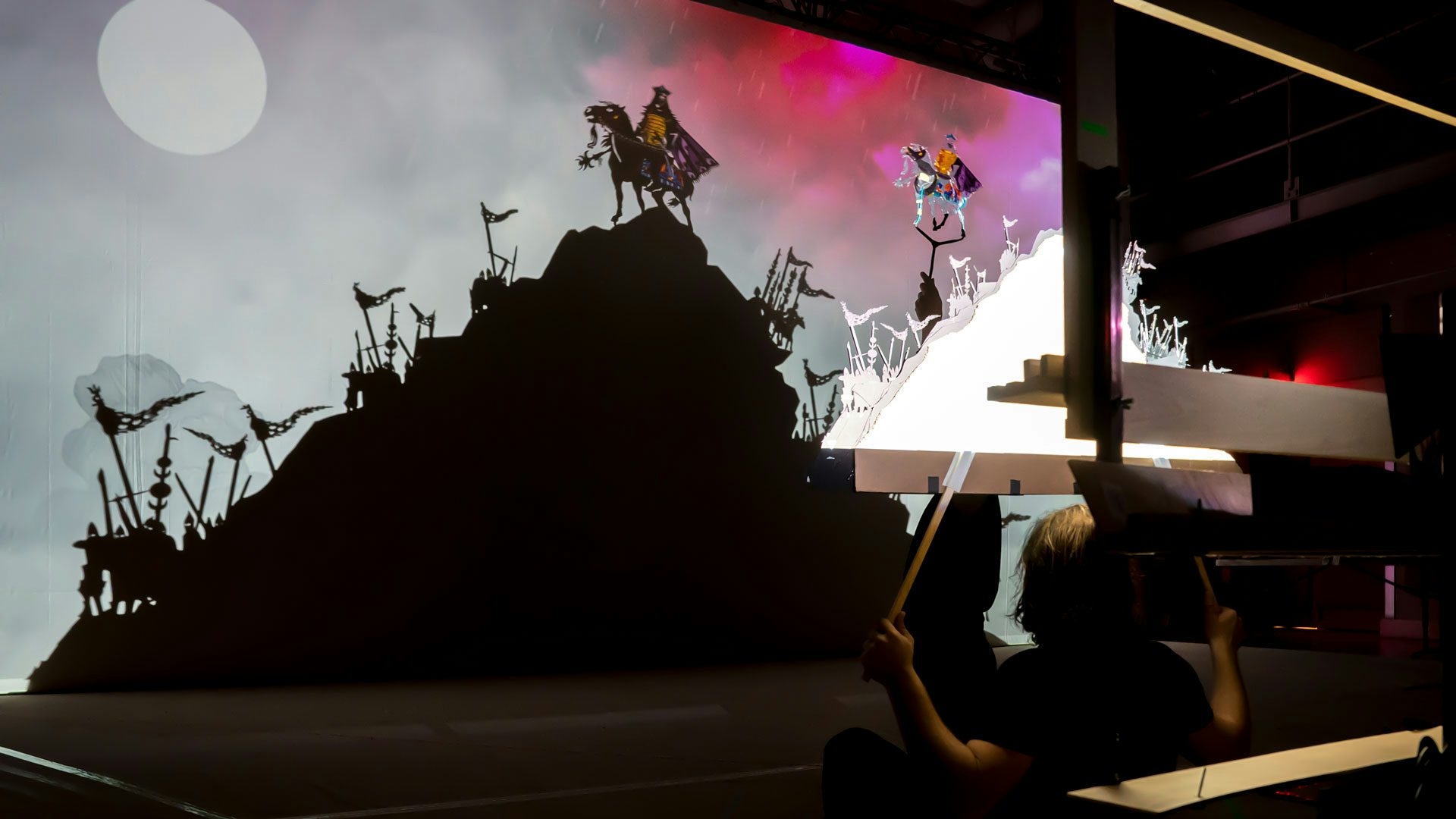 A person holds a prop of a mountain with soldiers along its sides against a light to cast a large shadow on a screen.