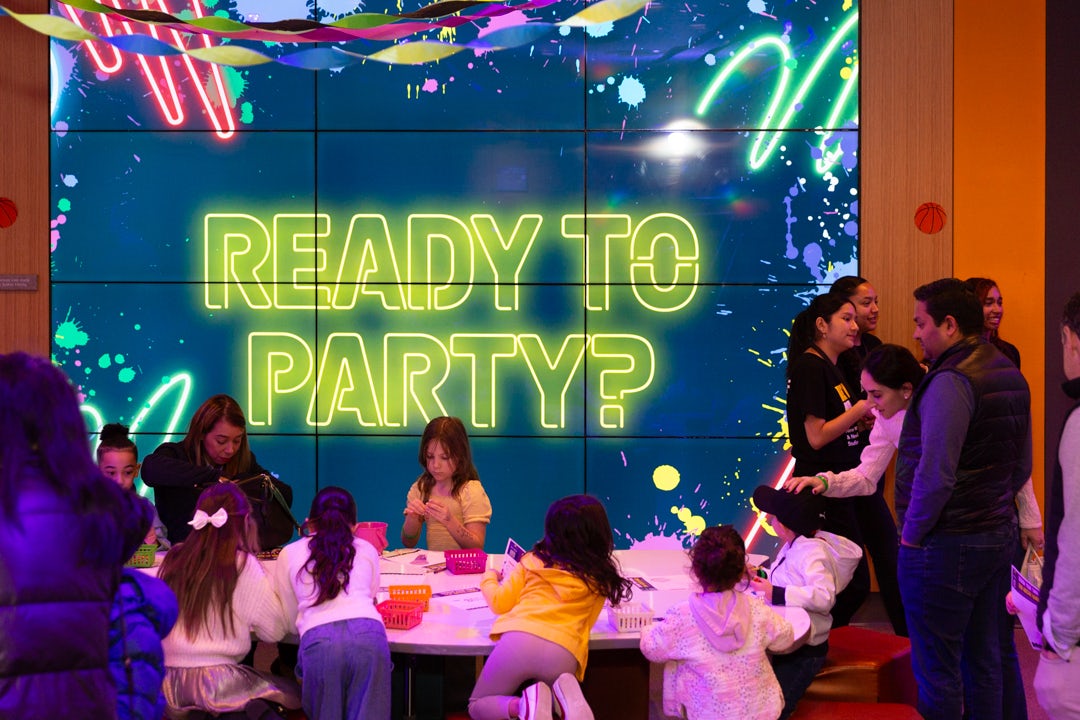 A group of kids and adults participate in pre-show activities in the New Victory Theater lower lobby in front a screen with yellow, neon text that reads "Ready to Party?"