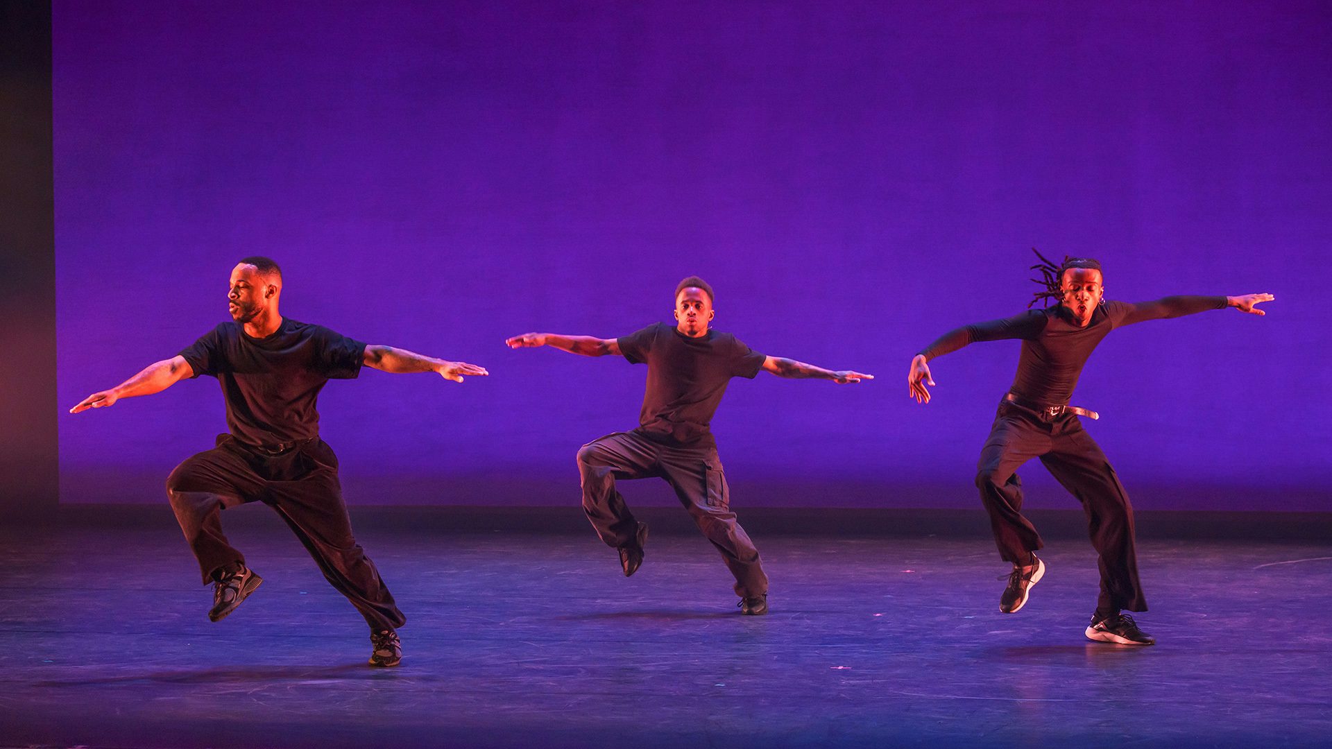 Three dancers wearing black clothes hold their arms out while jumping from side to side against an indigo background.
