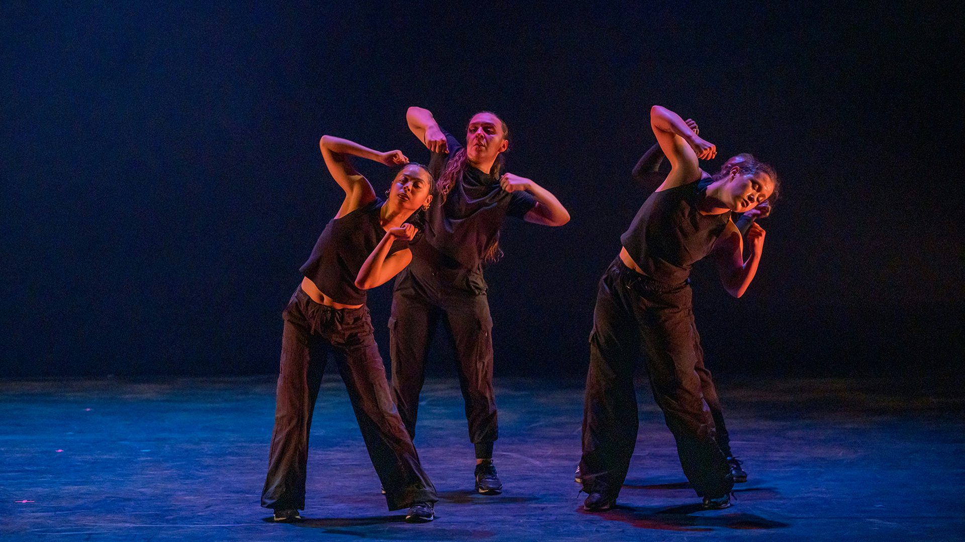 Four dancers wearing black clothes lean to the right with their arms up and bent near their faces against a black background.